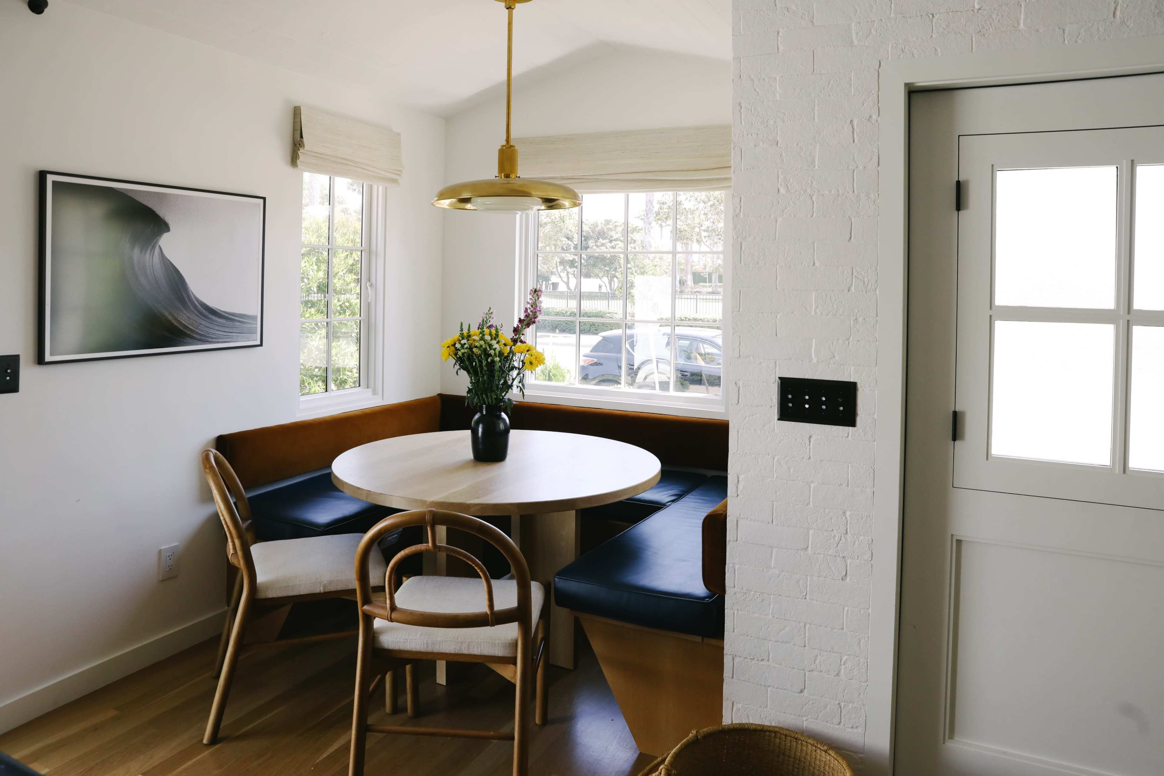 A dining nook features a round table surrounded by wooden chairs and a flower vase, with large windows providing natural light.