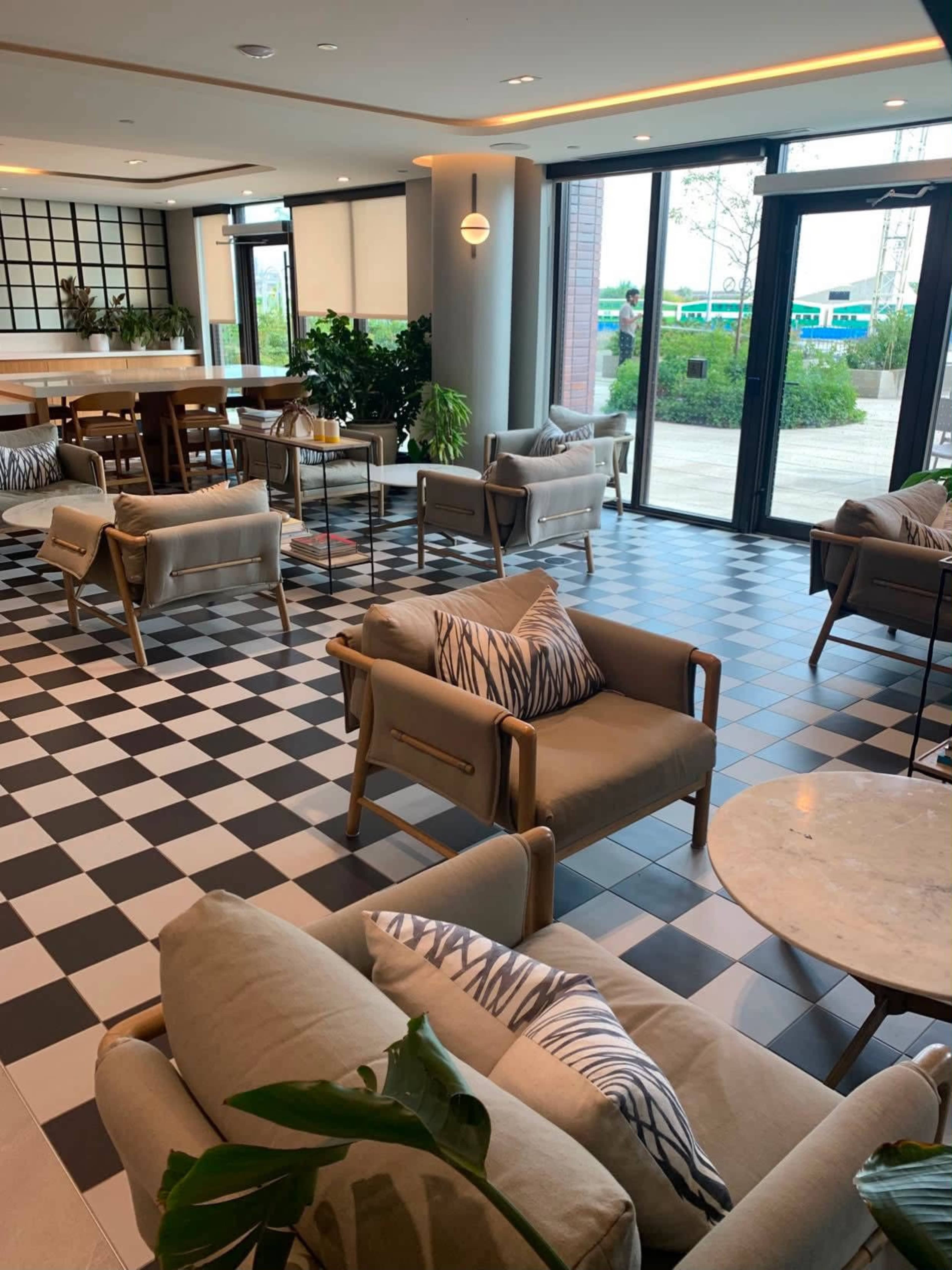 The image shows a modern lounge area with light-colored seating, checkered flooring, and large windows opening to an outdoor view.