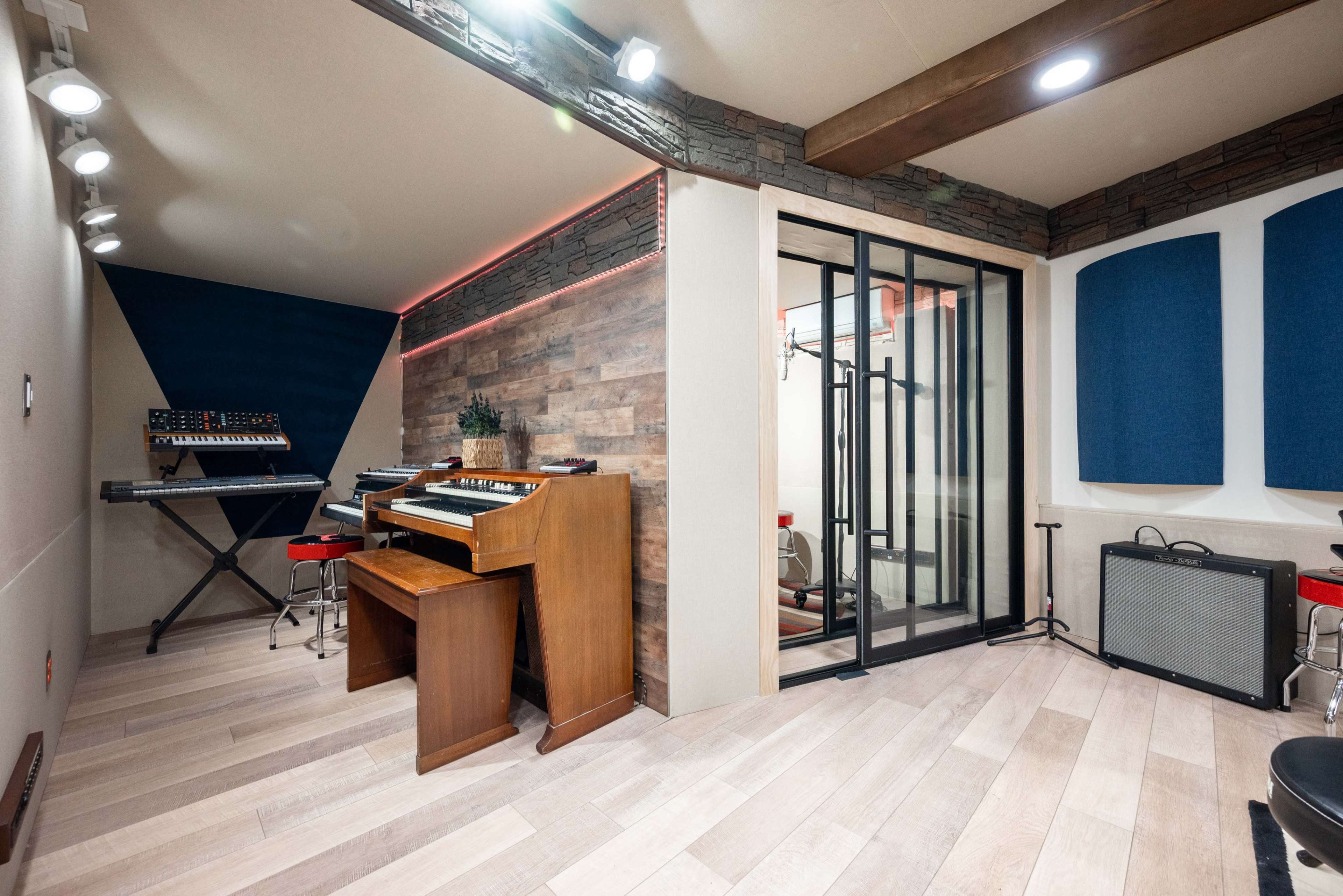 A modern music studio featuring a wooden keyboard, synthesizers, and soundproofing panels, with a glass enclosure for vocal recording.