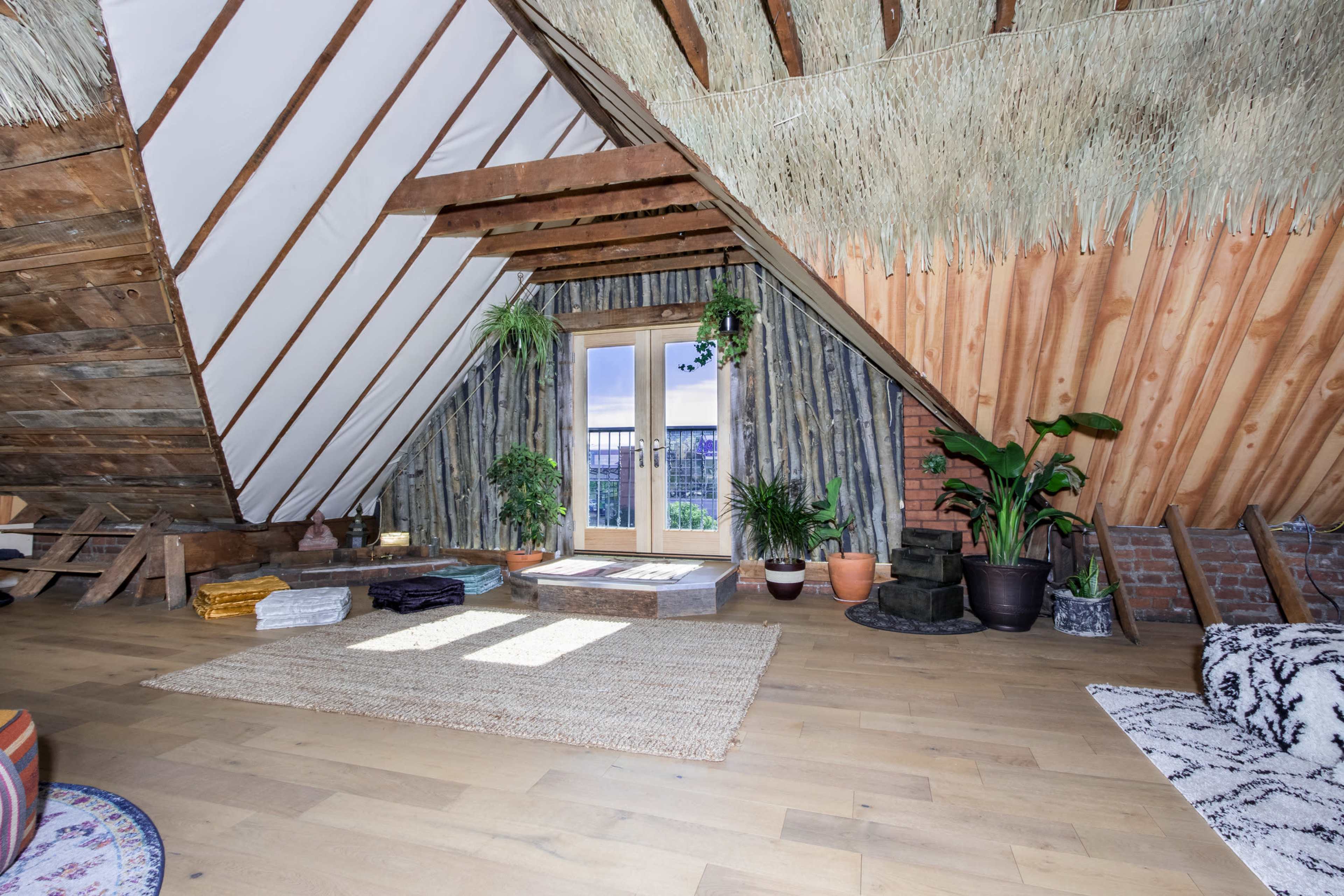A cozy attic space featuring wooden beams, a door leading to a balcony, and various potted plants on the floor and walls.