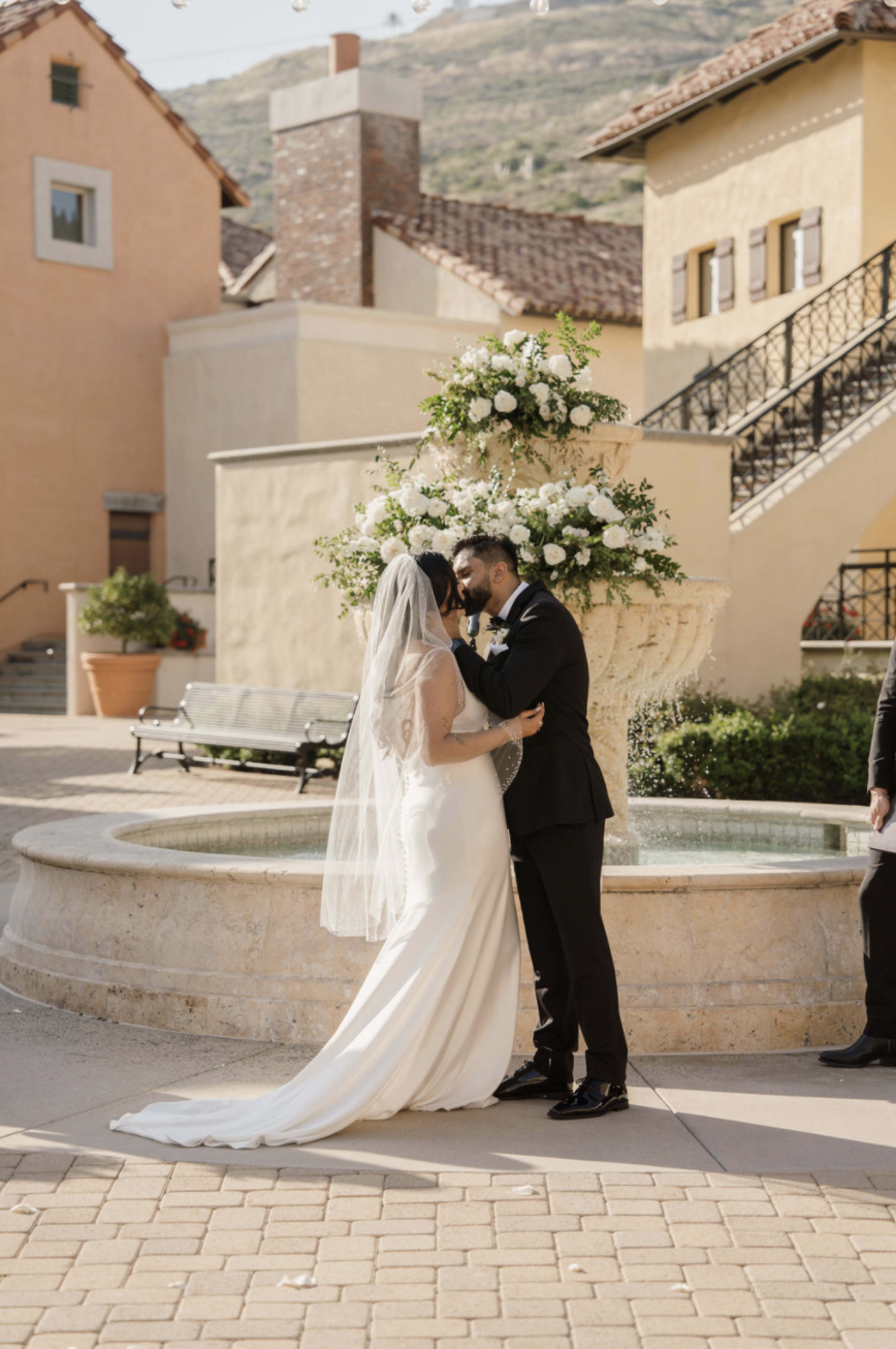 A couple exchanges vows in front of a floral arrangement by a fountain in an outdoor setting.
