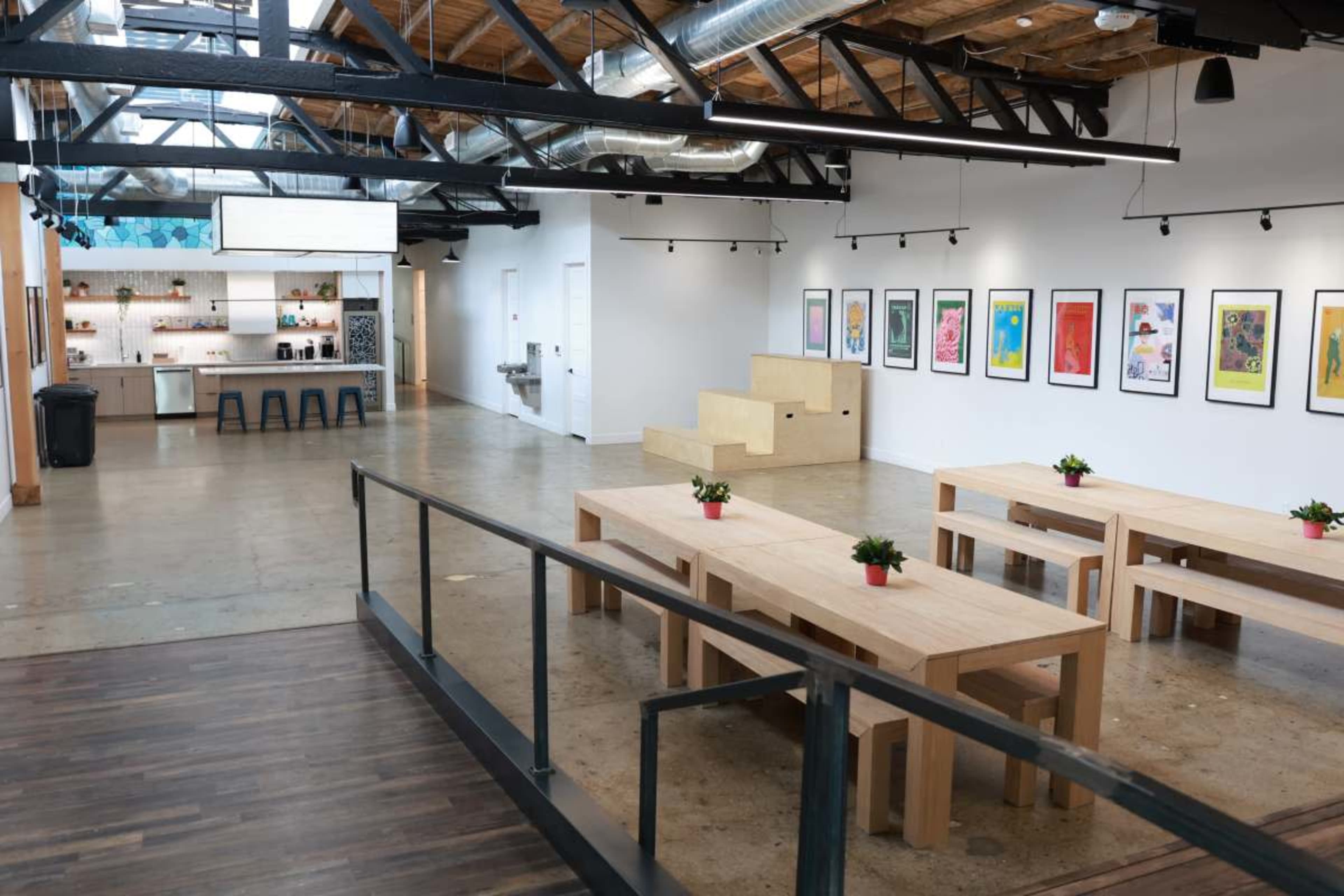 The image shows an open, modern workspace featuring wooden tables, colorful artwork on the walls, and a kitchen area in the background.