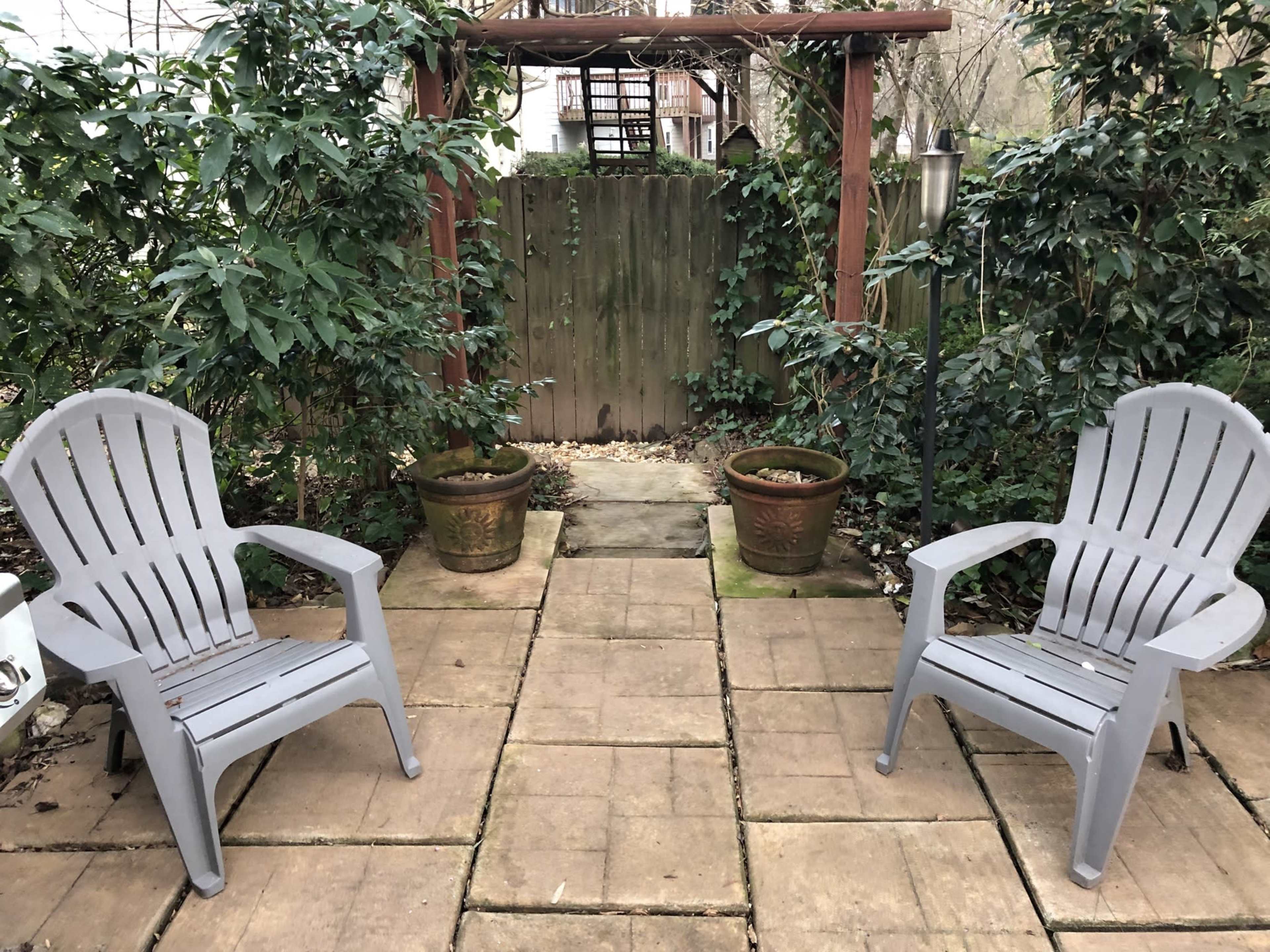 Two gray chairs are positioned on a stone patio surrounded by greenery and two potted plants.