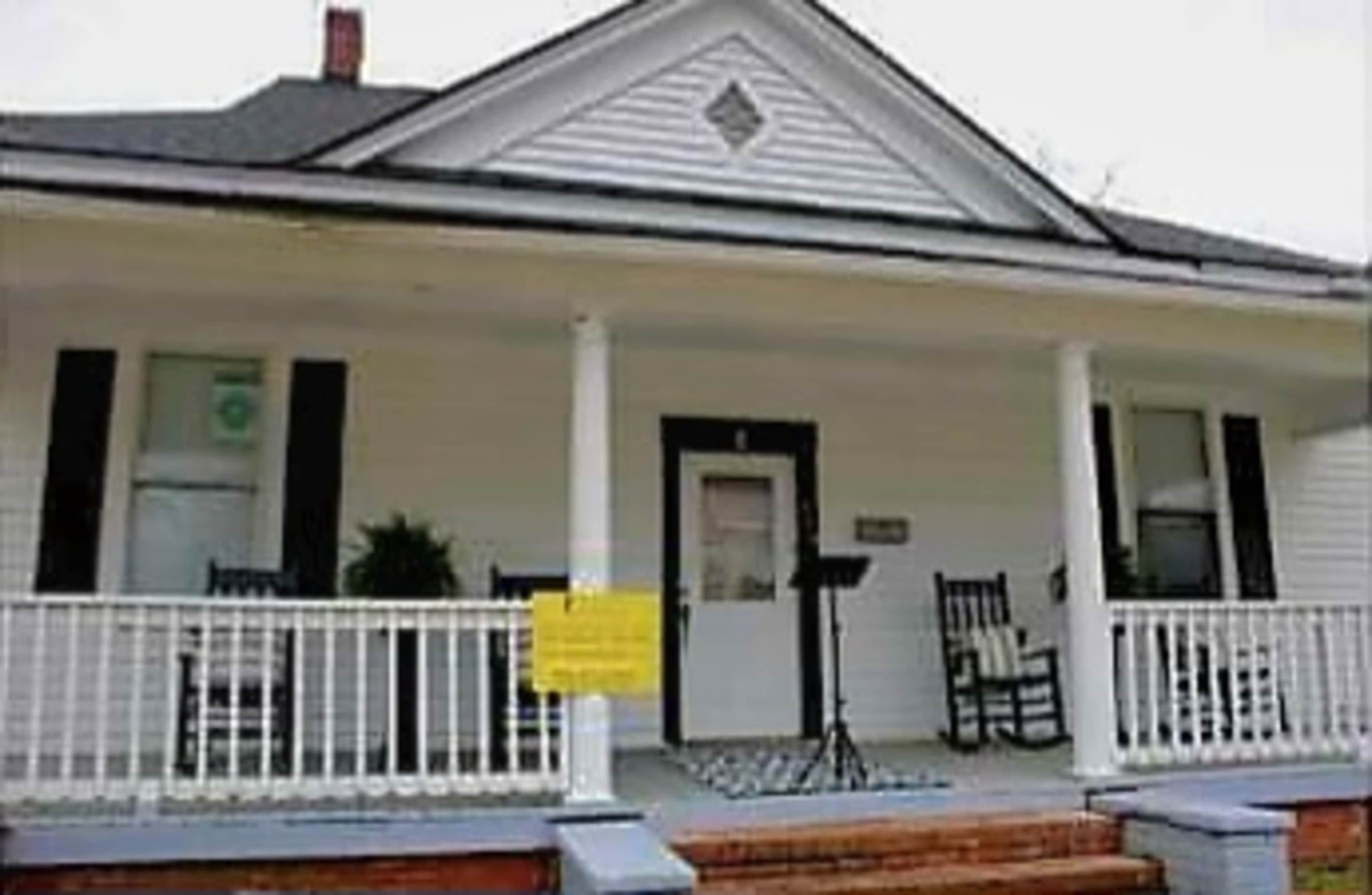 A white house with a covered porch, featuring rocking chairs and a sign on the railing.