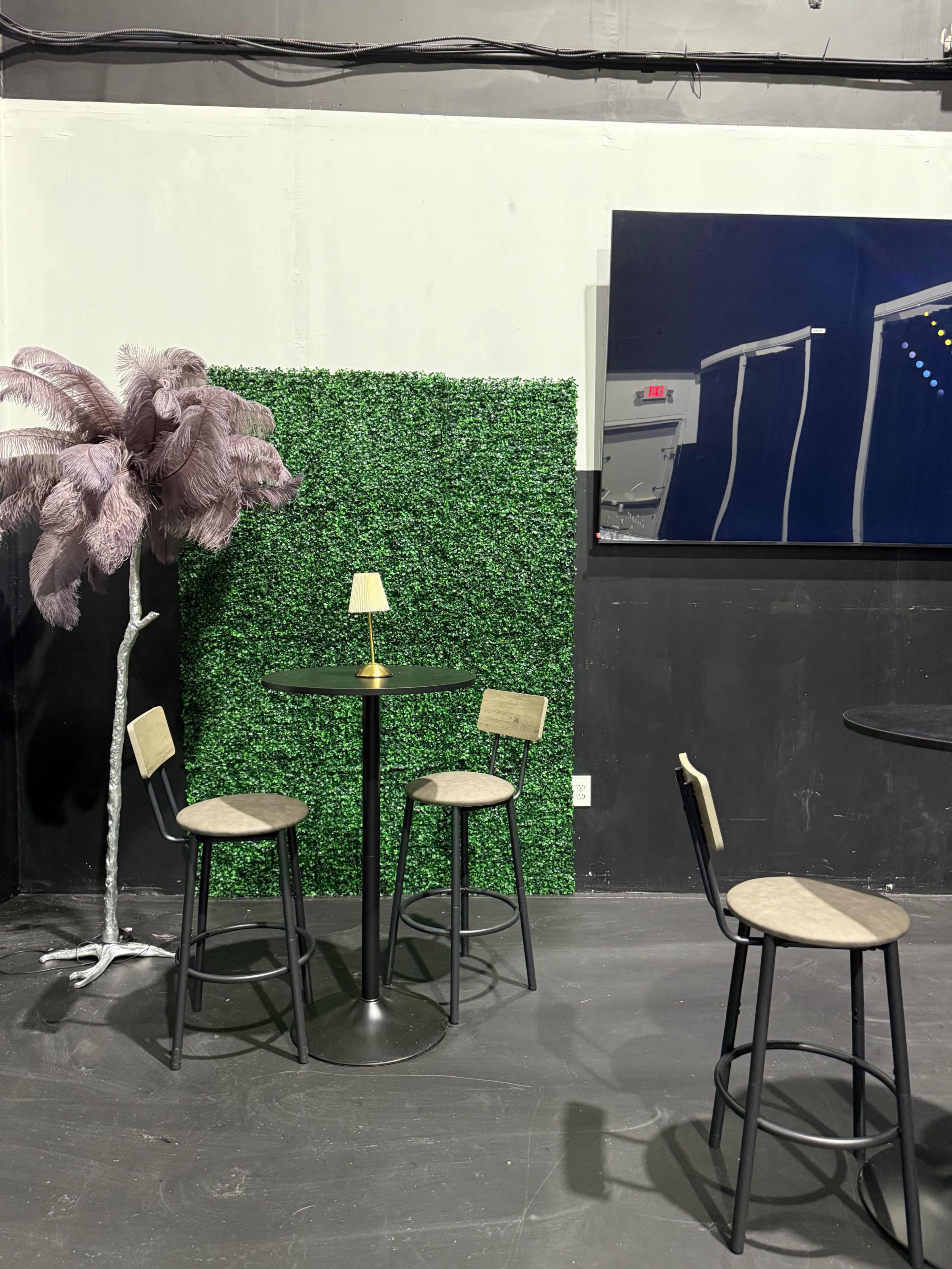 A small, round black table with four stools is set against a green artificial hedge and a large artwork on a dark wall.