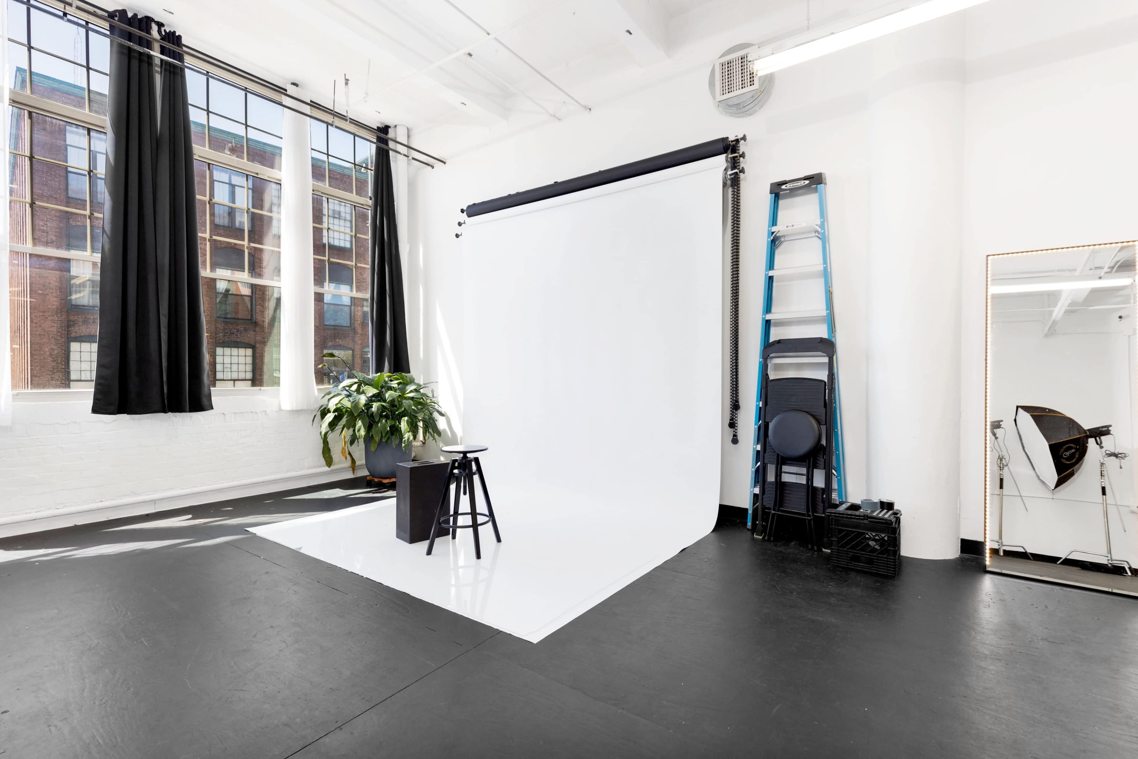 A well-lit photography studio featuring a white backdrop, a stool, a ladder, and a large potted plant, with black curtains framing the windows.