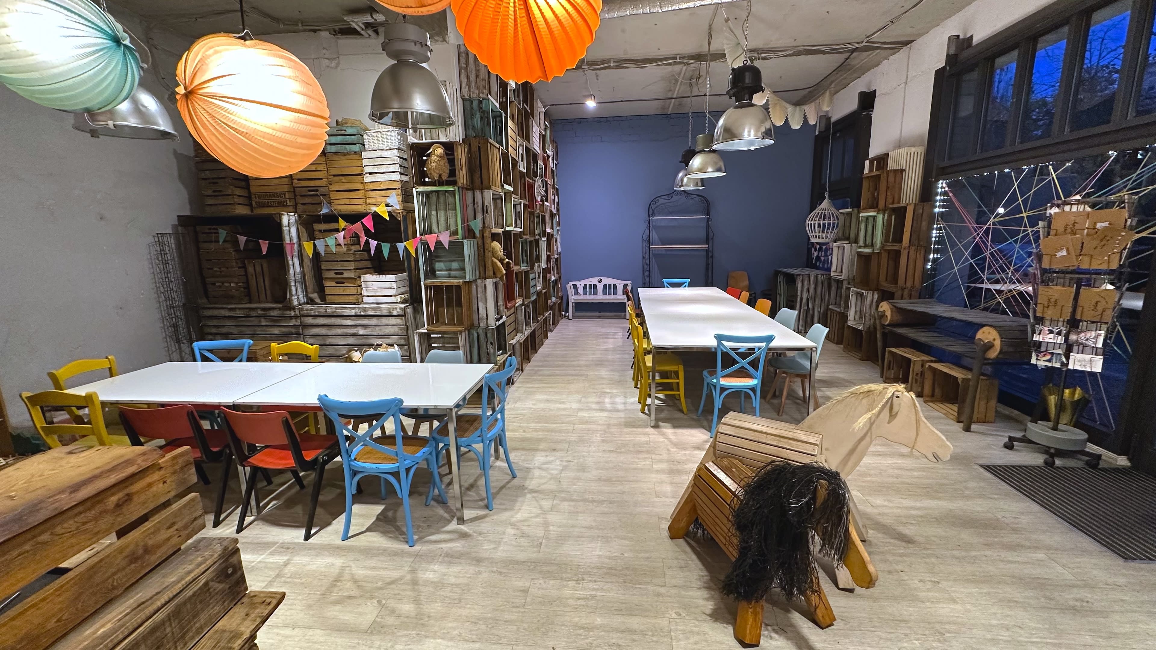 The image shows a spacious room filled with colorful chairs and tables, surrounded by wooden crates stacked on shelves and hanging lanterns.