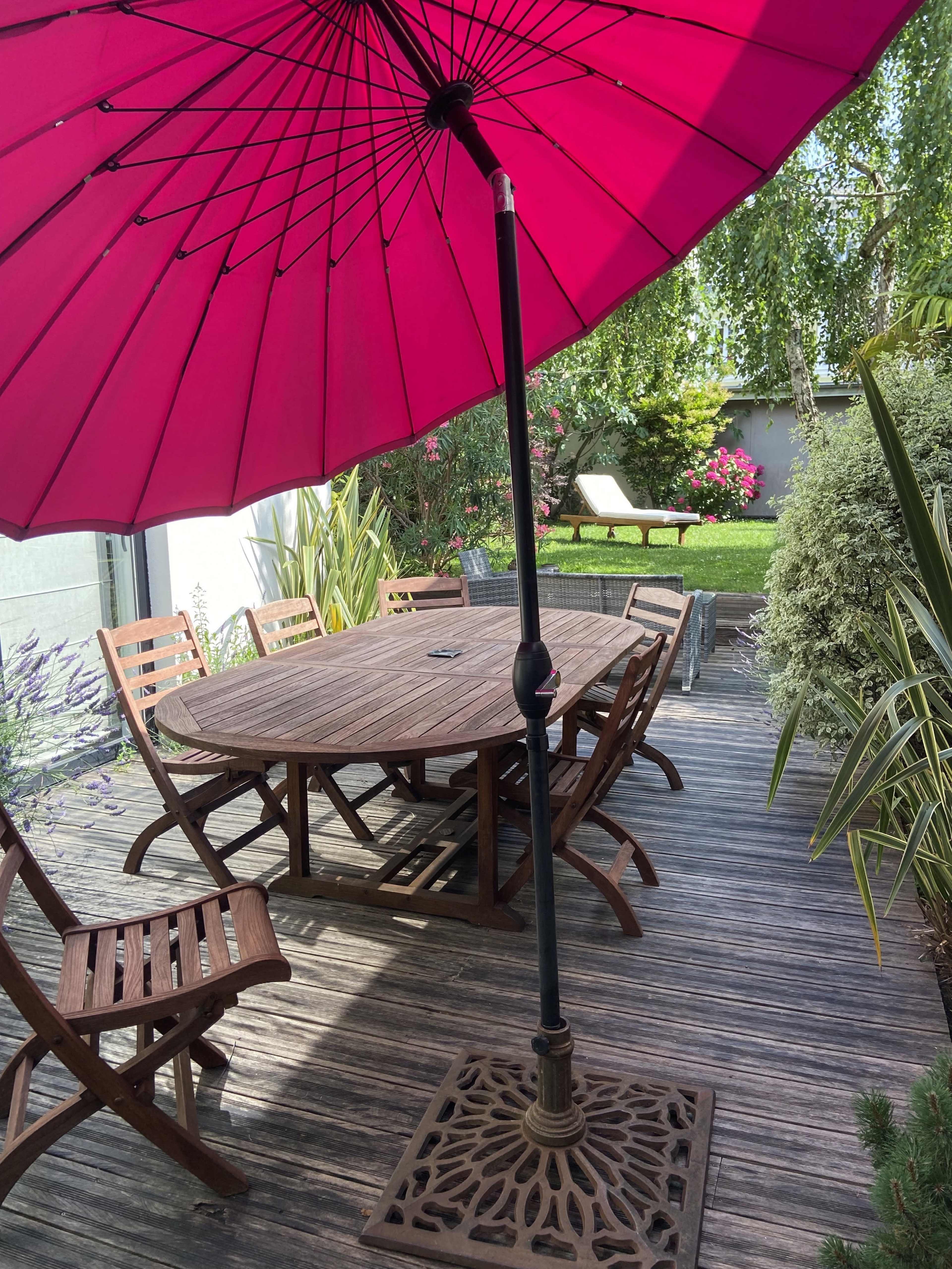 A pink patio umbrella stands over a wooden table surrounded by chairs on a deck with greenery and a lawn in the background.