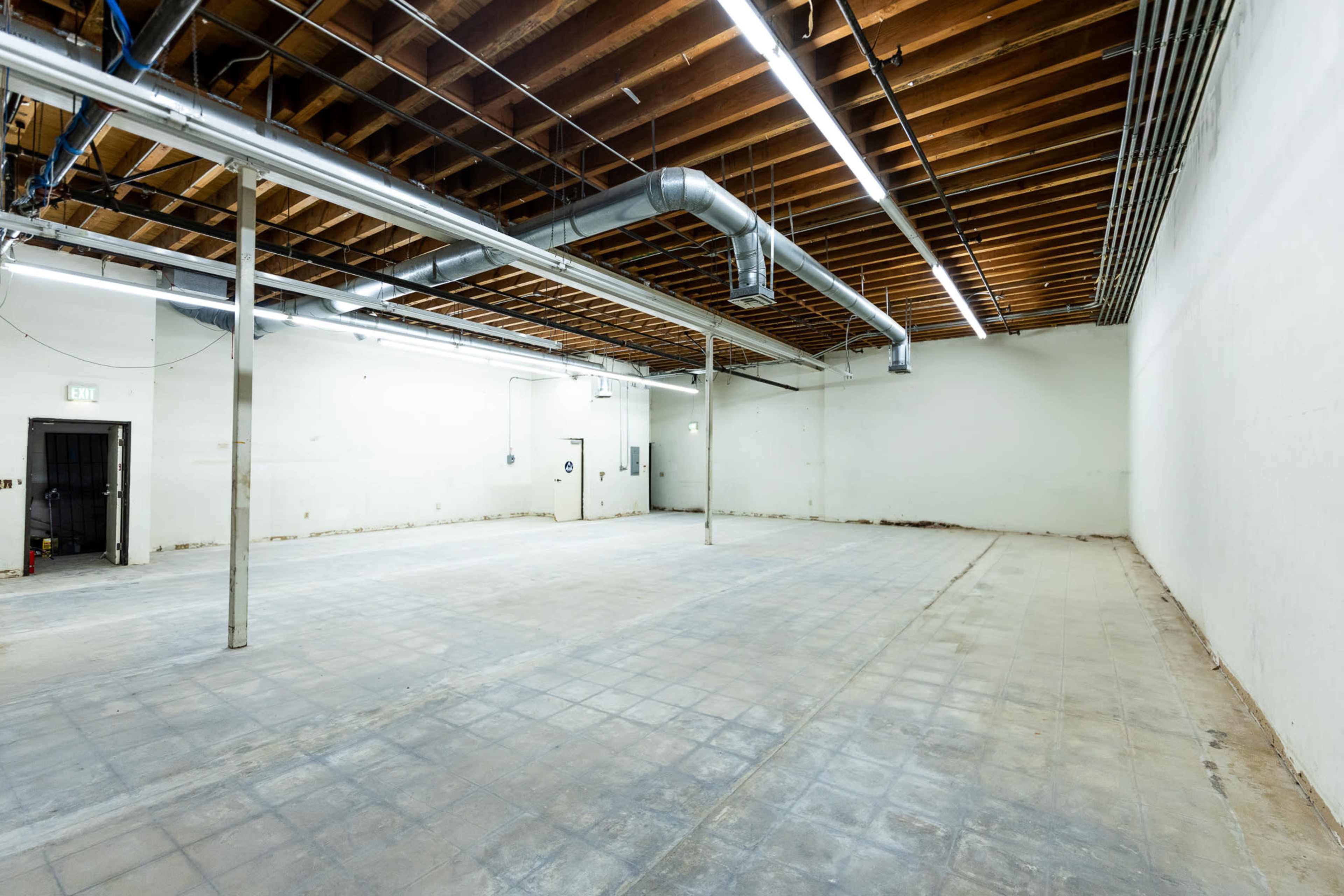 The image shows a large, empty warehouse space with a concrete floor, exposed wooden beams, and ventilation ducts overhead.