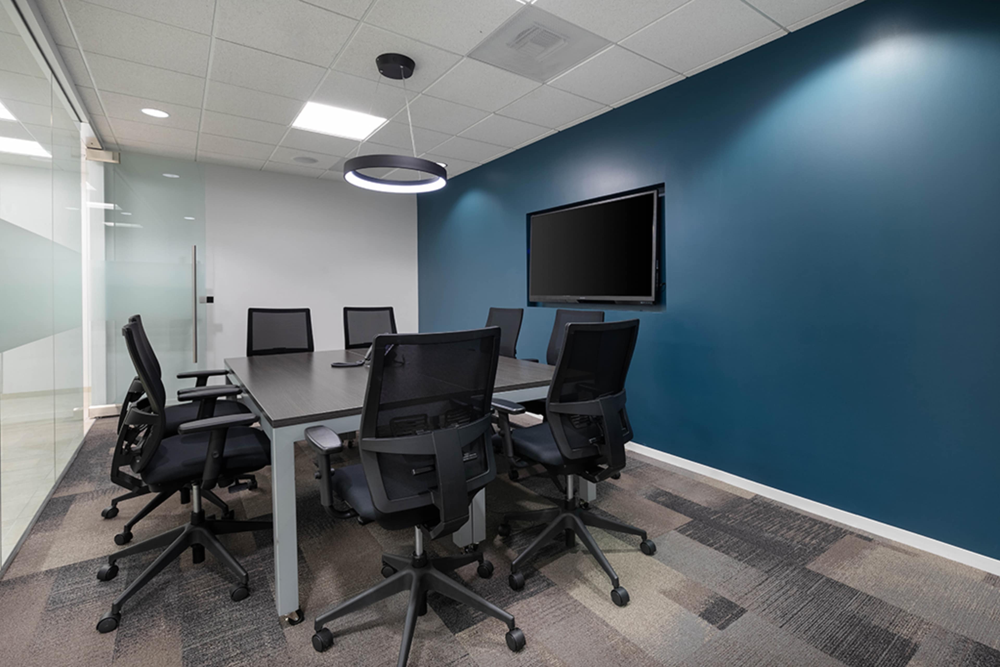 A modern conference room features a large table surrounded by ergonomic chairs, with a mounted TV and a teal accent wall.