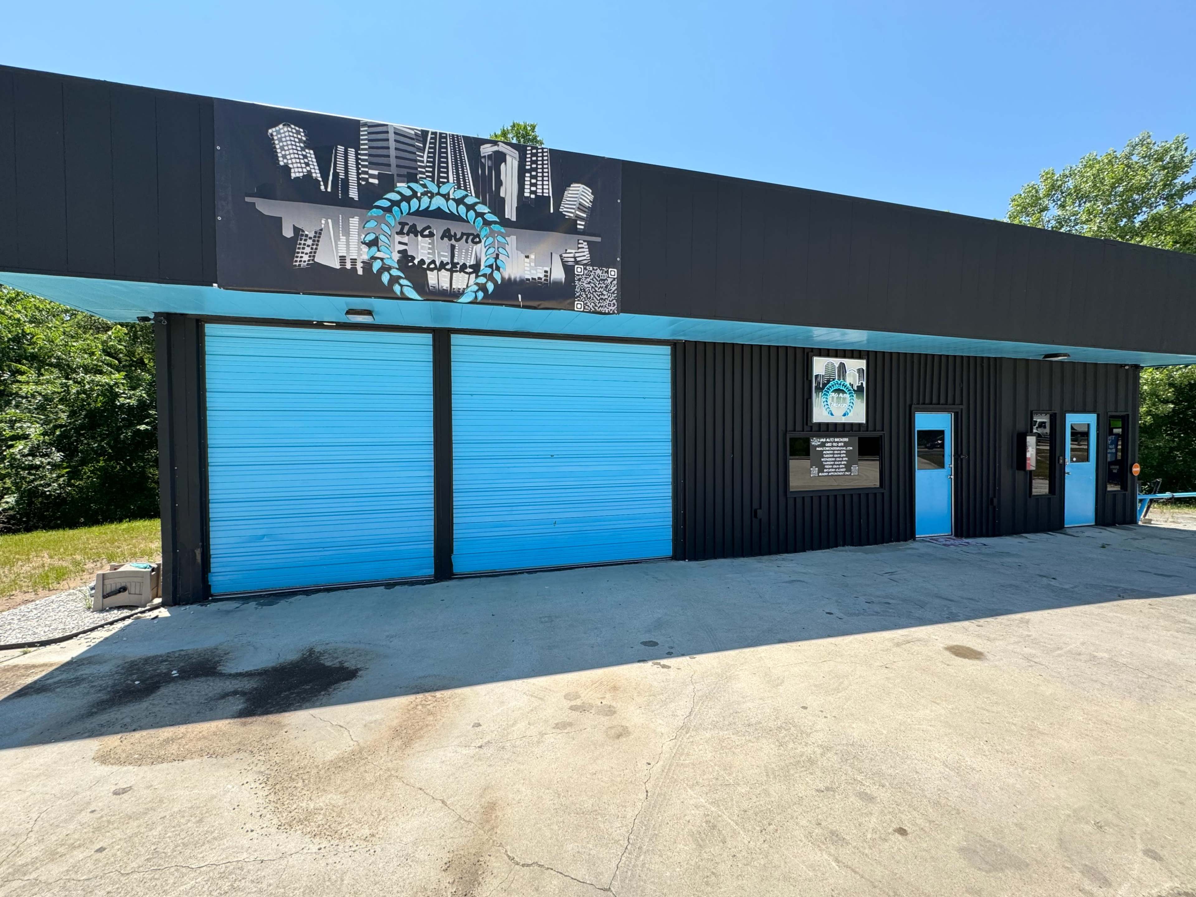 The image shows a car repair shop with a black and blue exterior, featuring two large blue roll-up doors and a sign above the entrance.