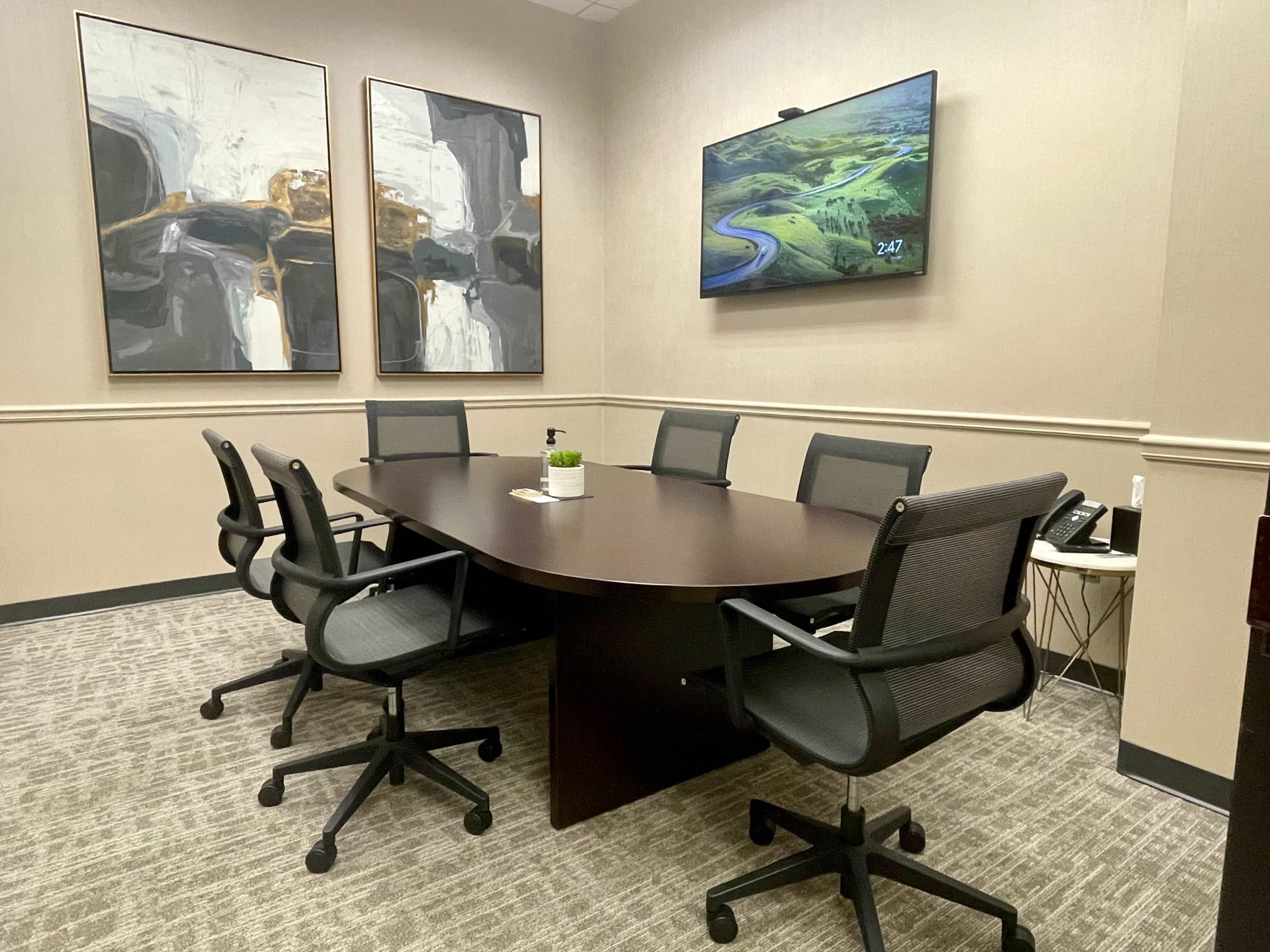 A conference room with a large oval table surrounded by six black office chairs, a television mounted on the wall, and abstract artwork on the adjacent walls.