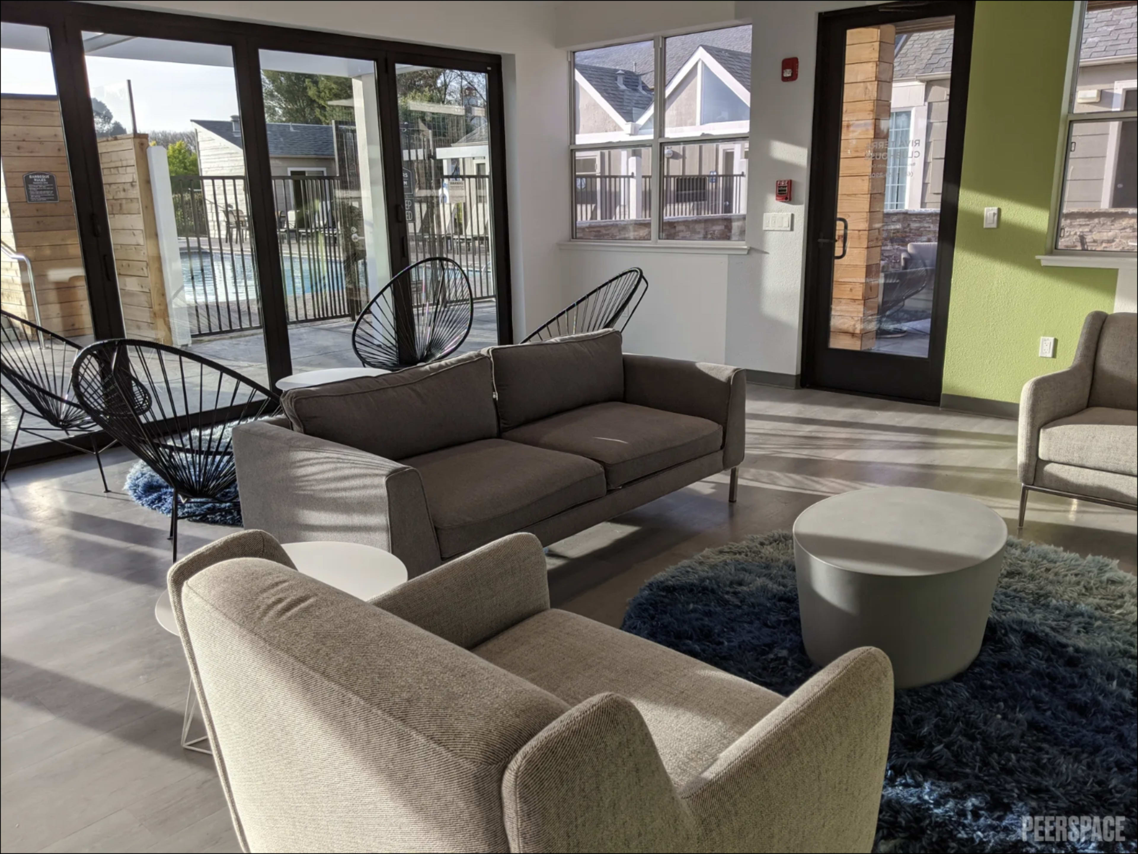 The image shows a modern common area with a seating arrangement of gray sofas and chairs, a round coffee table, and large windows revealing a pool outside.