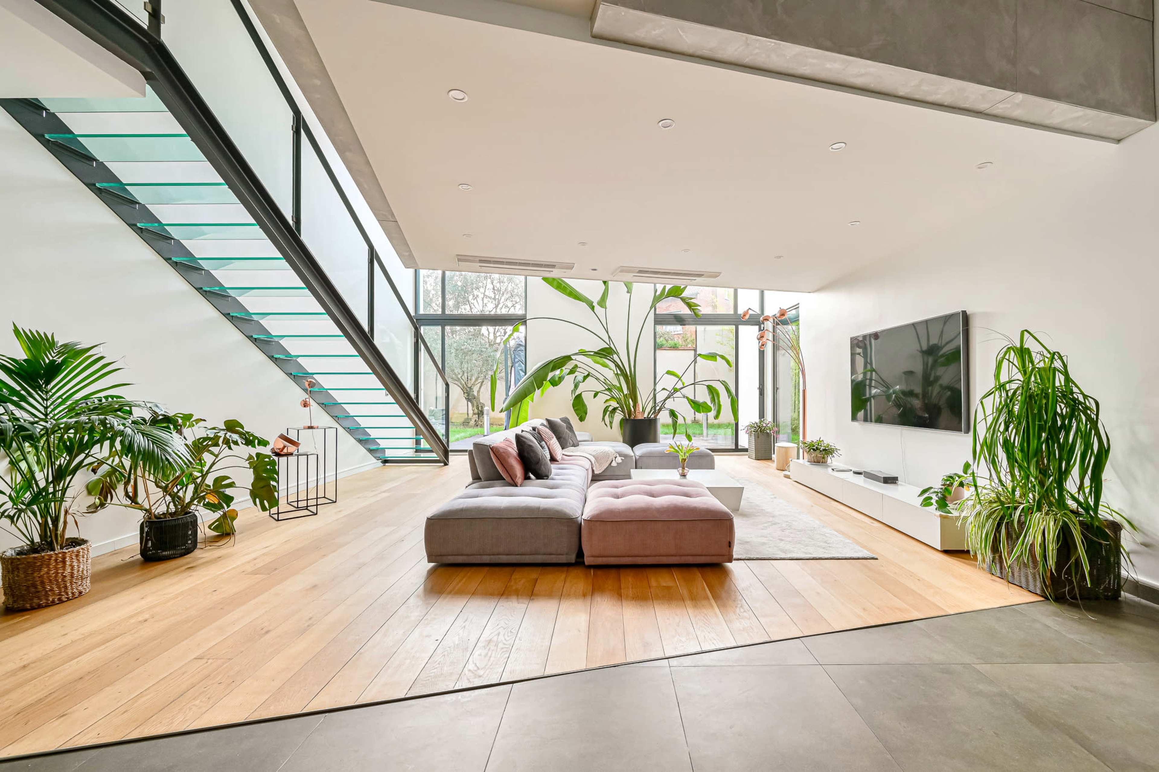 A modern living room features a large sectional sofa, a flat-screen TV mounted on a white wall, and tall windows with indoor plants surrounding a wooden floor.