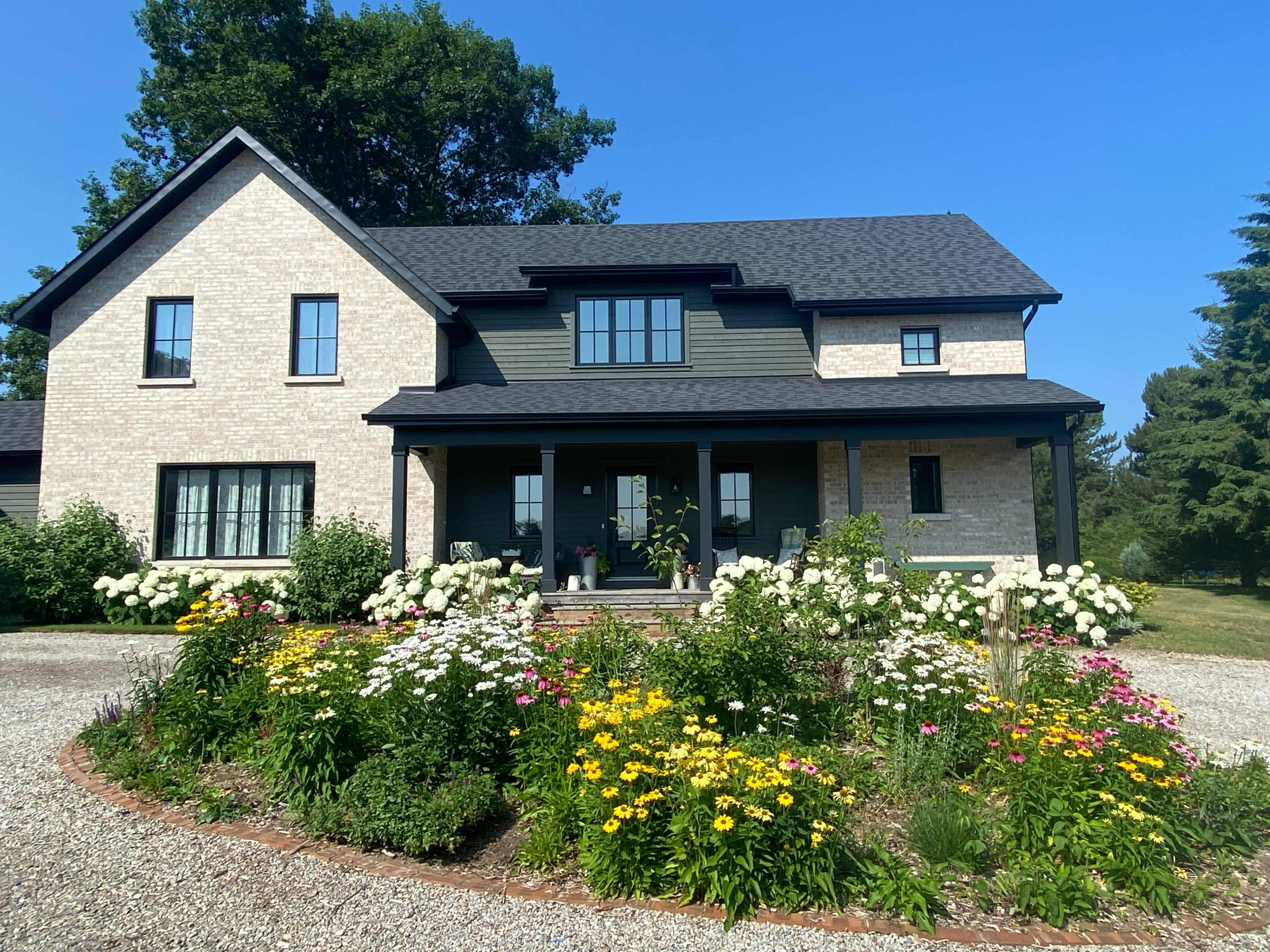 A two-story house with a mix of light brick and dark siding is set behind a landscaped garden featuring various colorful flowers and gravel pathways.