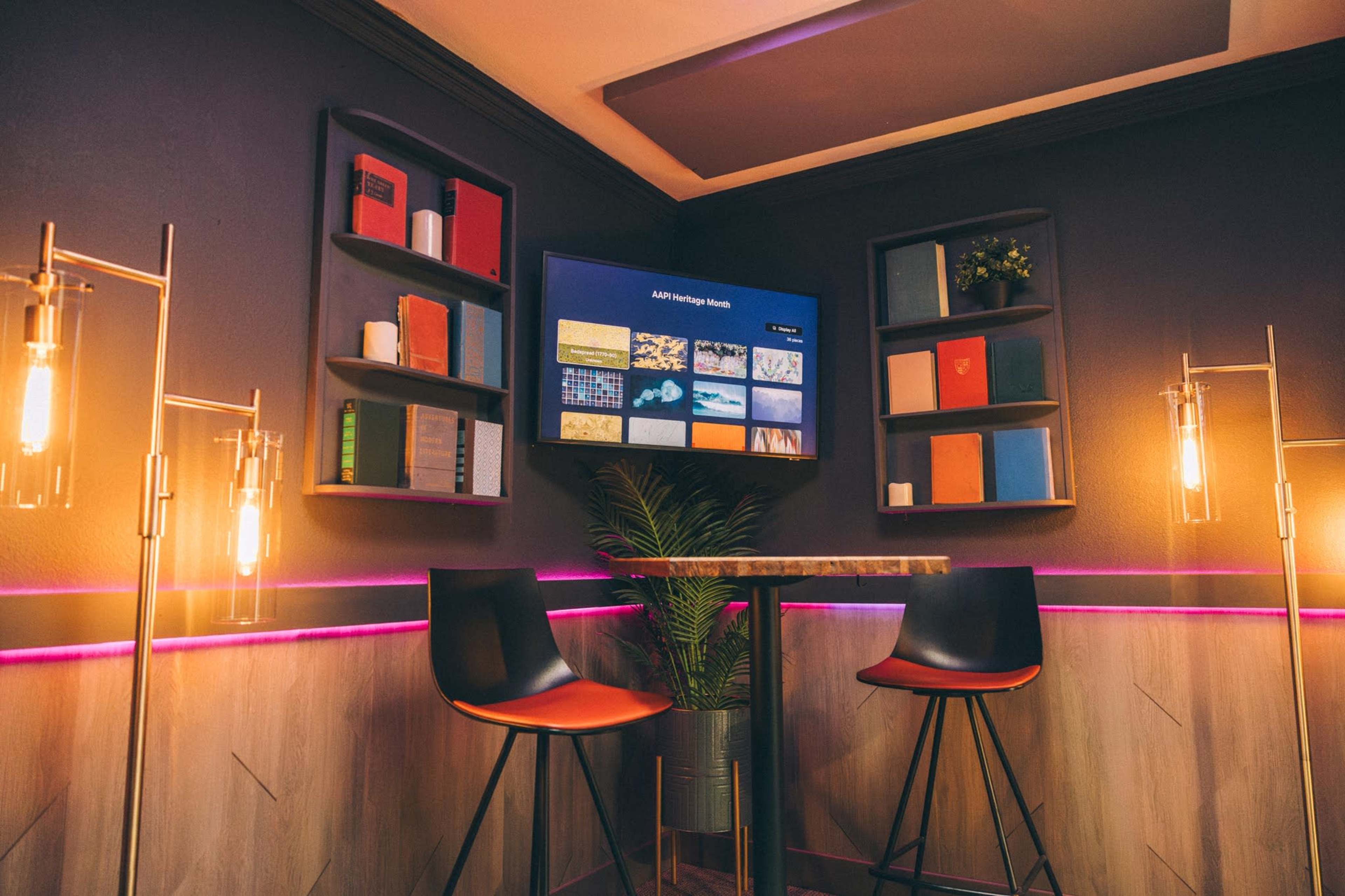 The image showcases a modern, cozy corner of a room featuring two high stools, a small table, a television screen, and decorative shelving with books and plants.