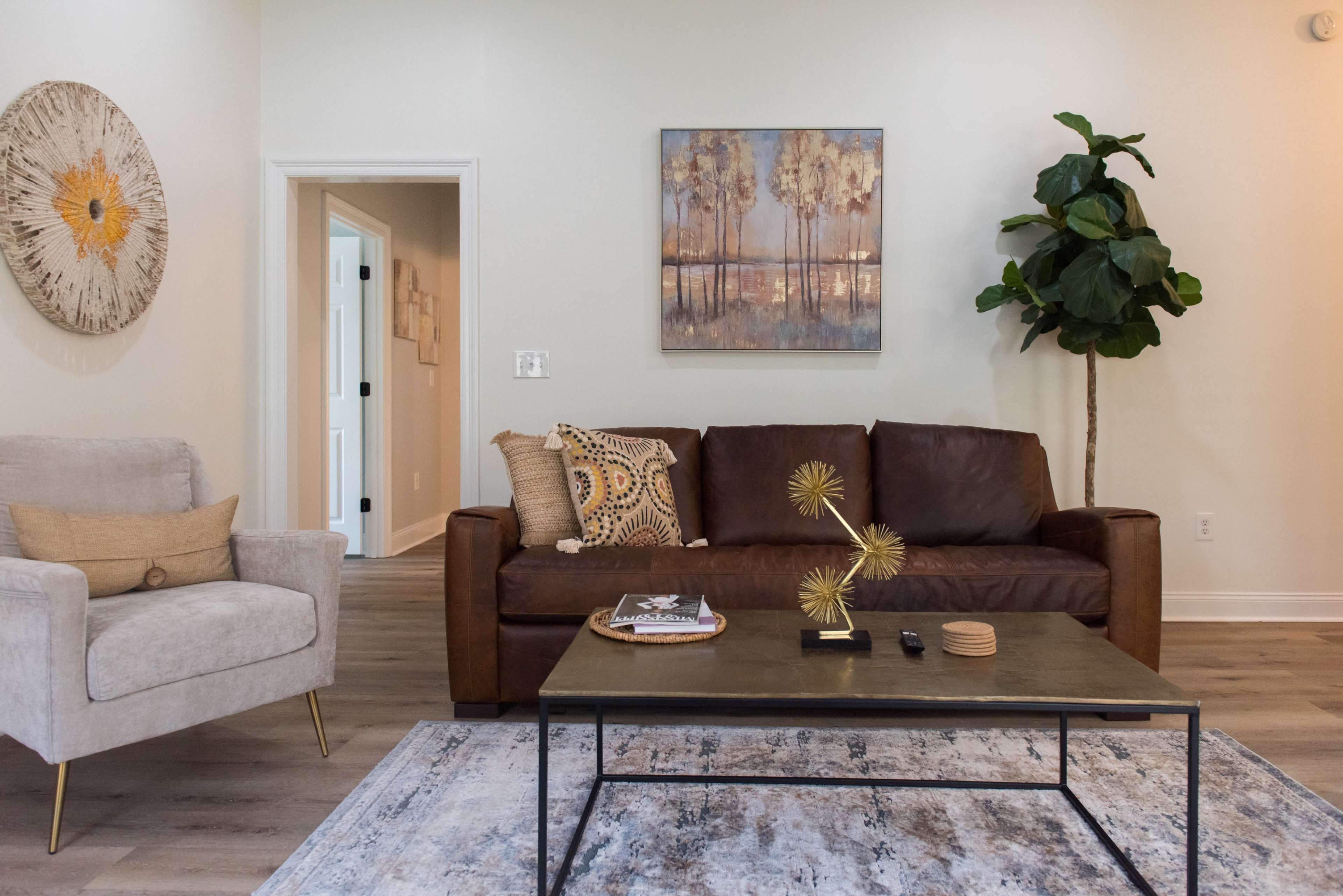 A brown leather sofa and a light gray armchair are arranged around a black coffee table on a patterned area rug in a well-lit living room featuring a wall painting and a potted plant.