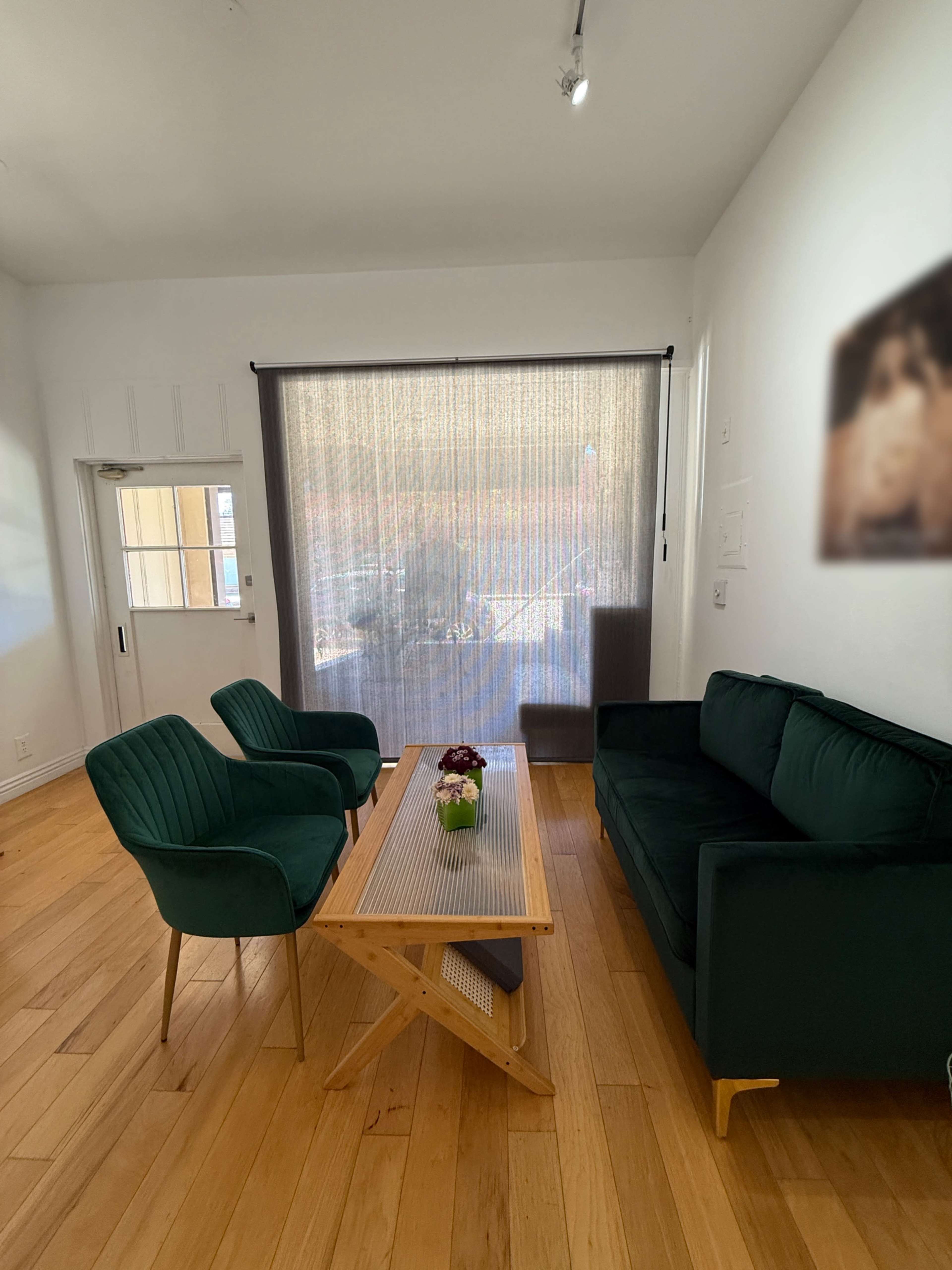 A living room features two green velvet chairs and a green couch around a wooden table, all positioned near a window covered by sheer curtains.