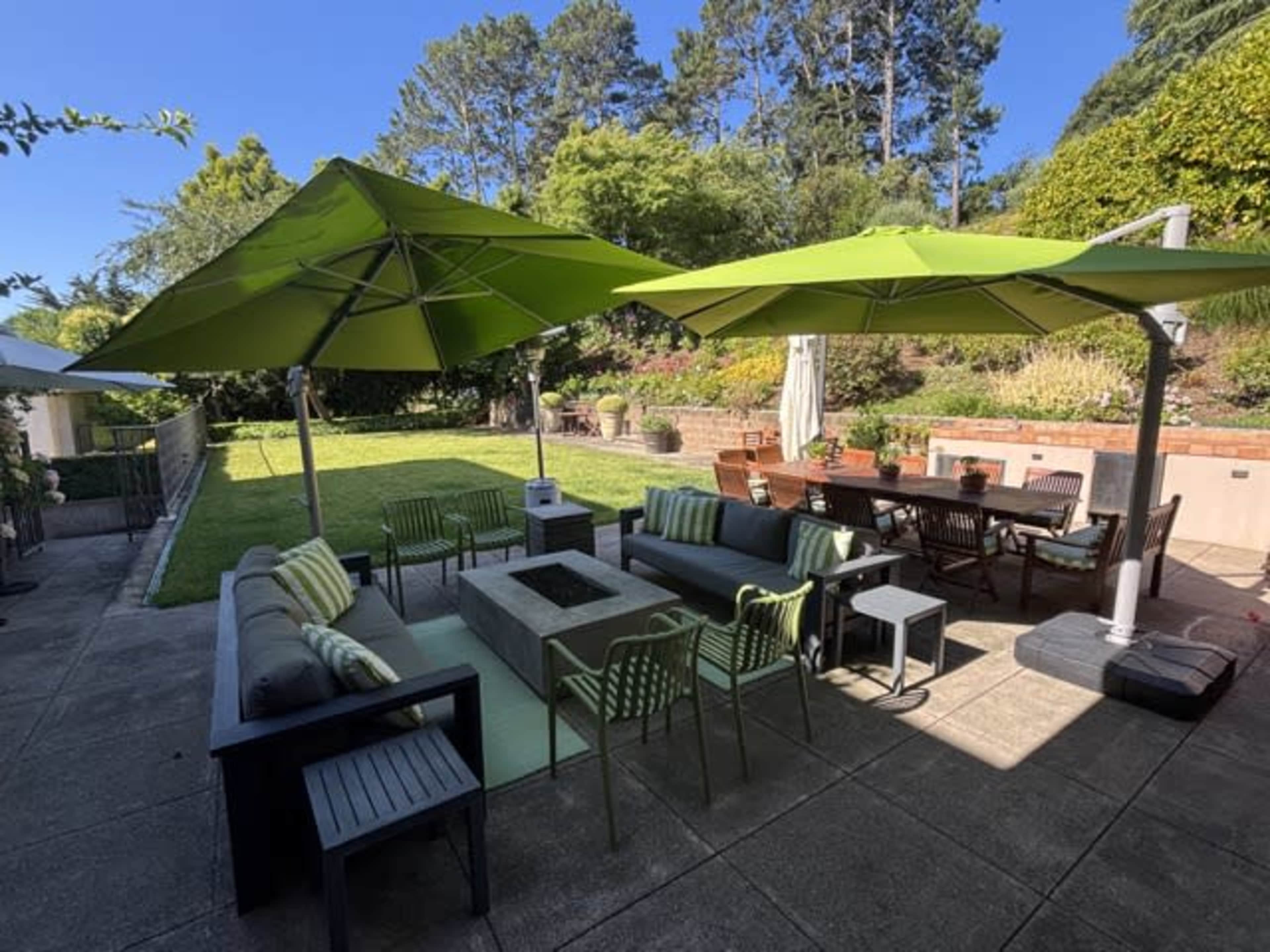 The image shows a well-arranged backyard with green umbrellas over seating areas and a dining table, surrounded by neatly maintained grass and garden features.