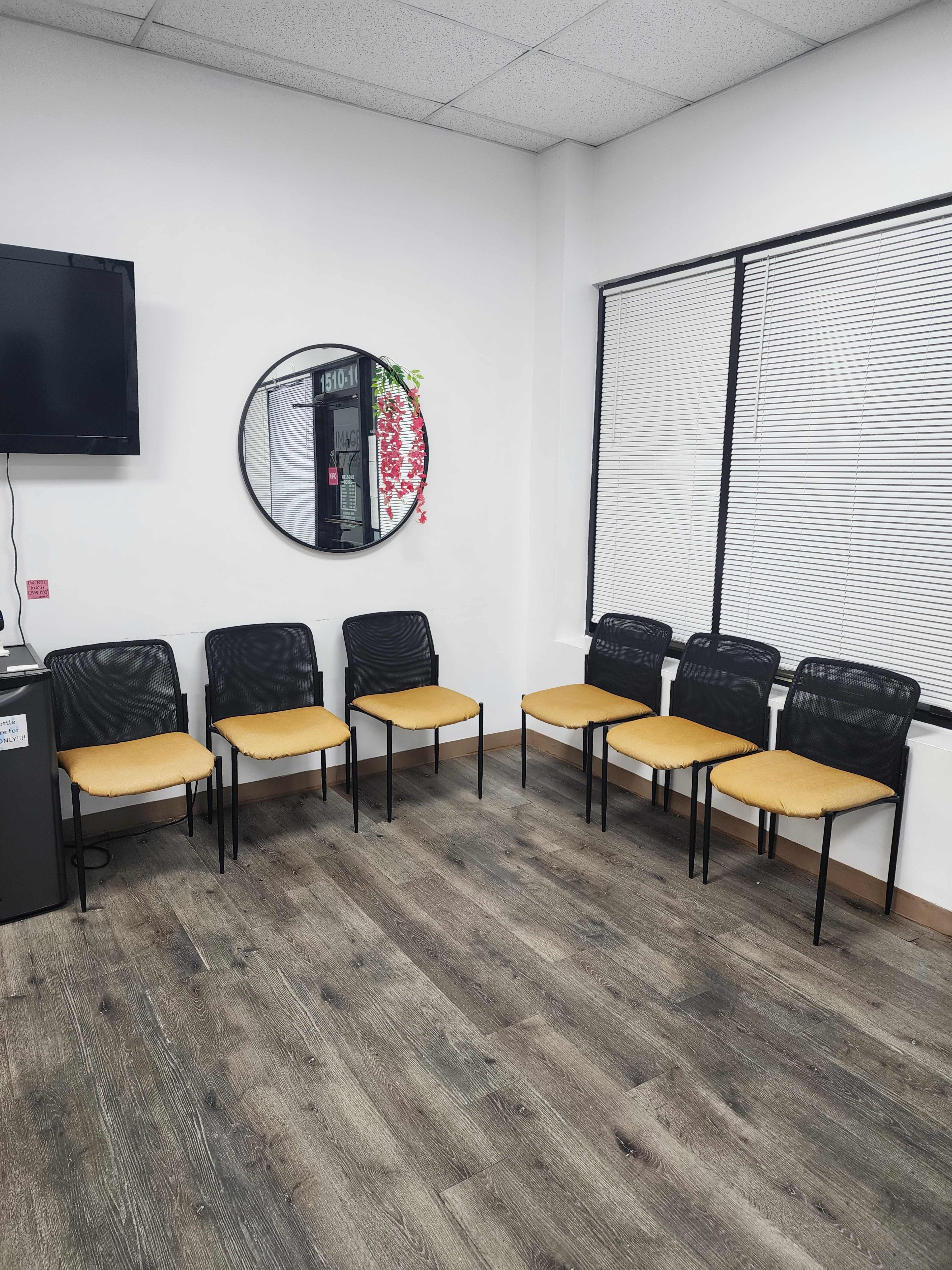 The image shows an empty waiting area with six black chairs and yellow seat cushions arranged against a wall with a round mirror and a television.