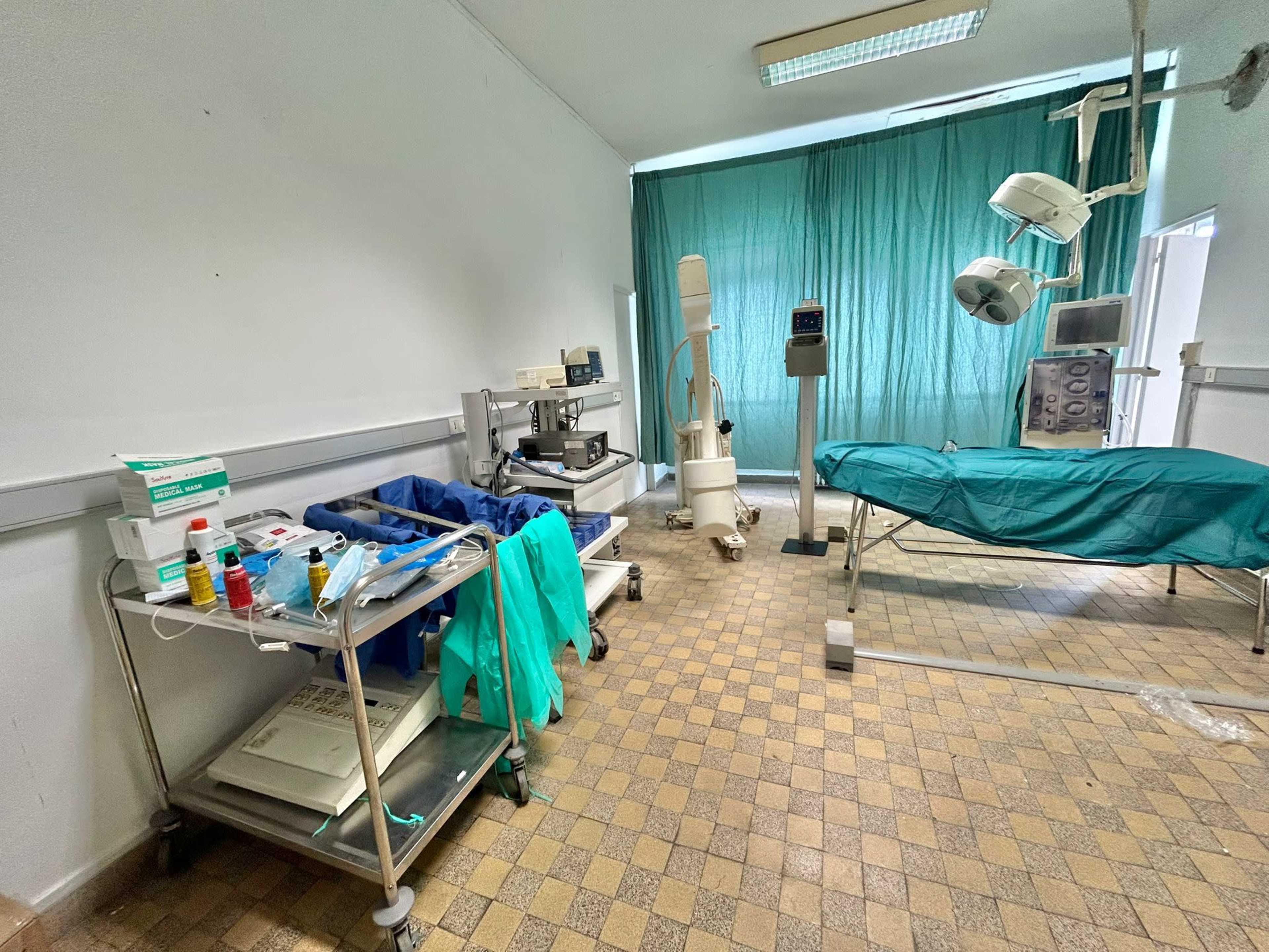 A medical room with a green surgical bed, medical equipment on carts, and curtains separating the space.