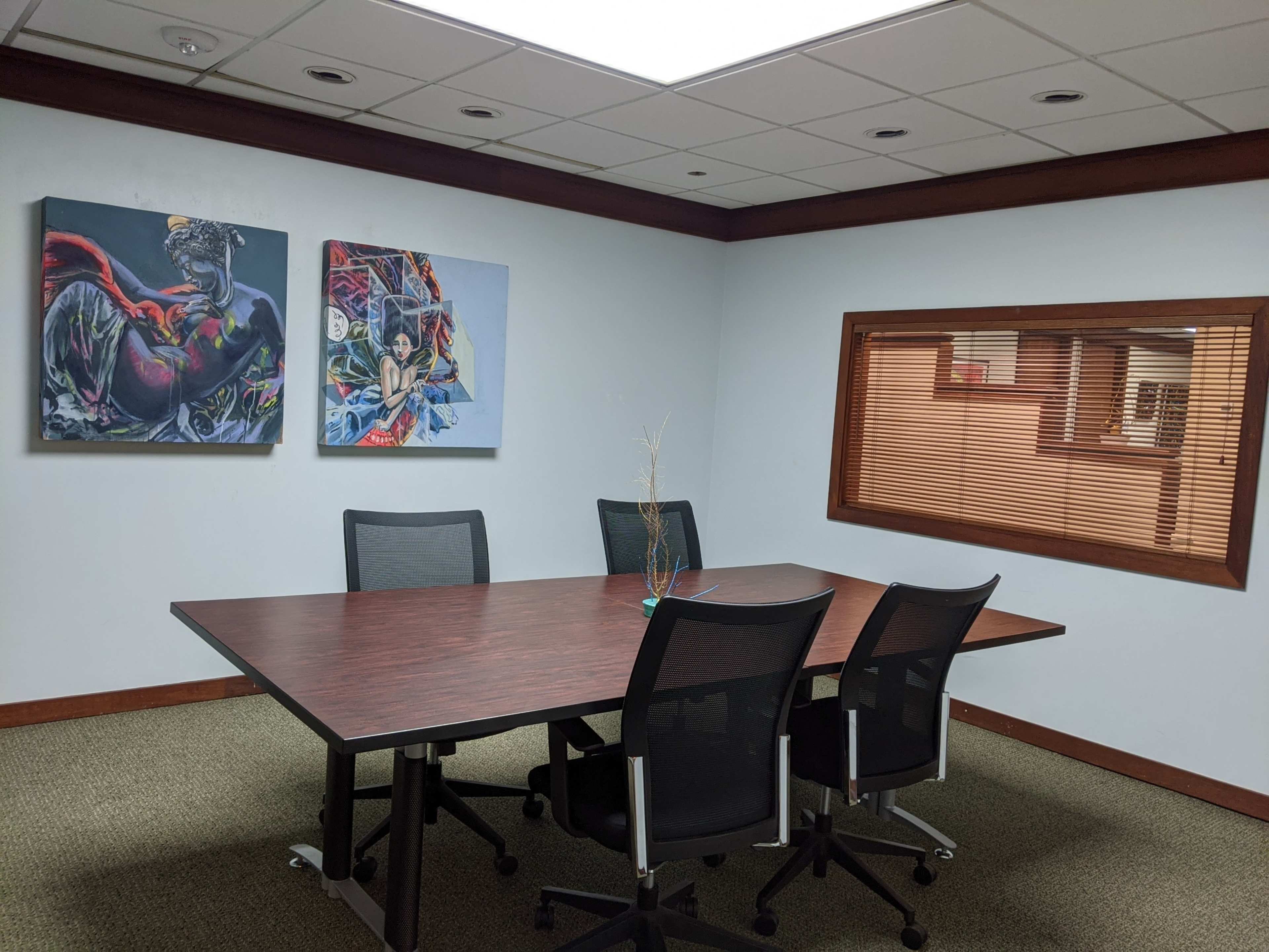 The image shows a conference room with a large wooden table surrounded by four black office chairs, and two colorful paintings hung on the wall opposite a window with blinds.