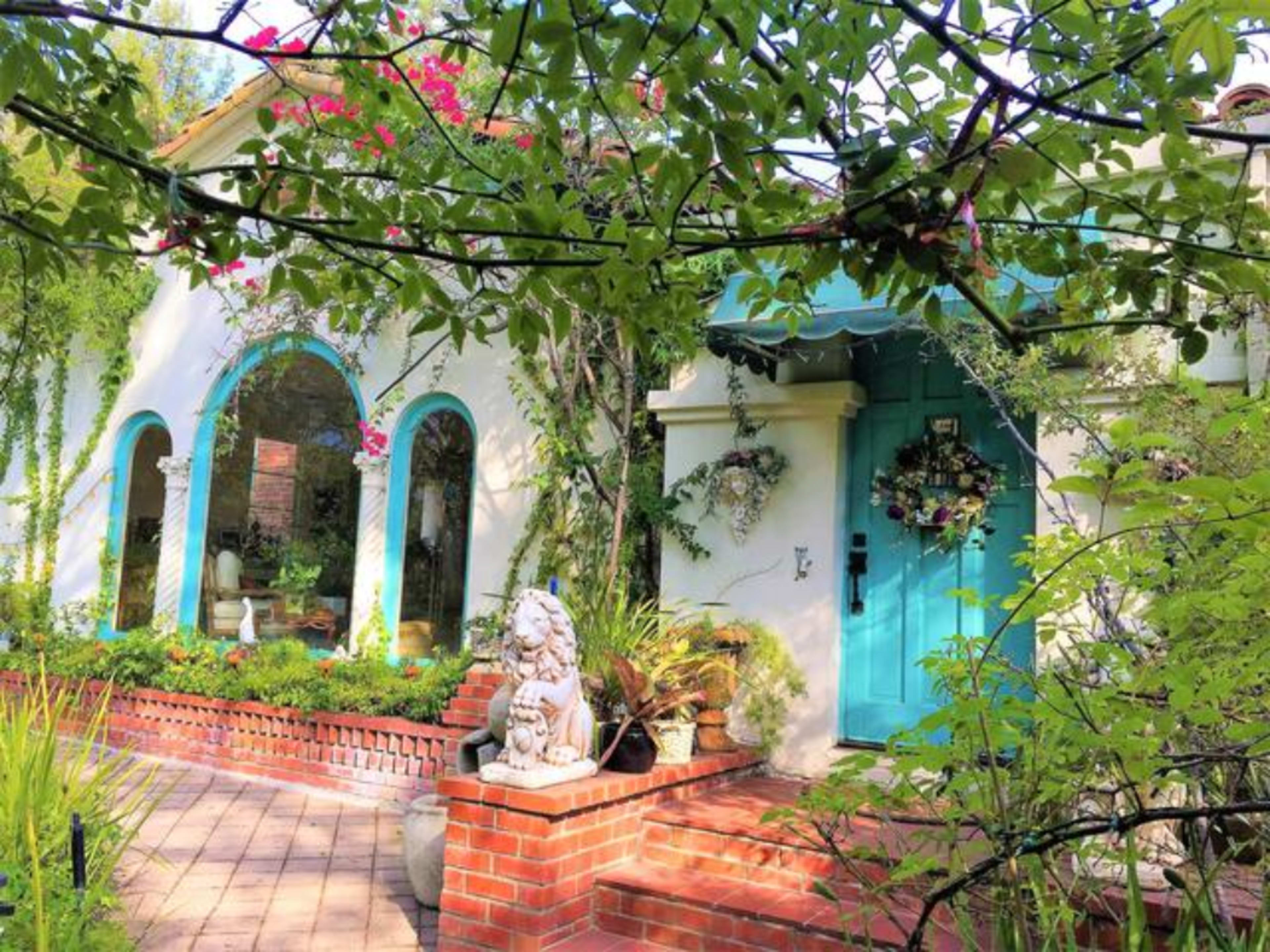 A house with an aqua door and arched windows is surrounded by greenery and features a stone lion statue on a brick pathway.