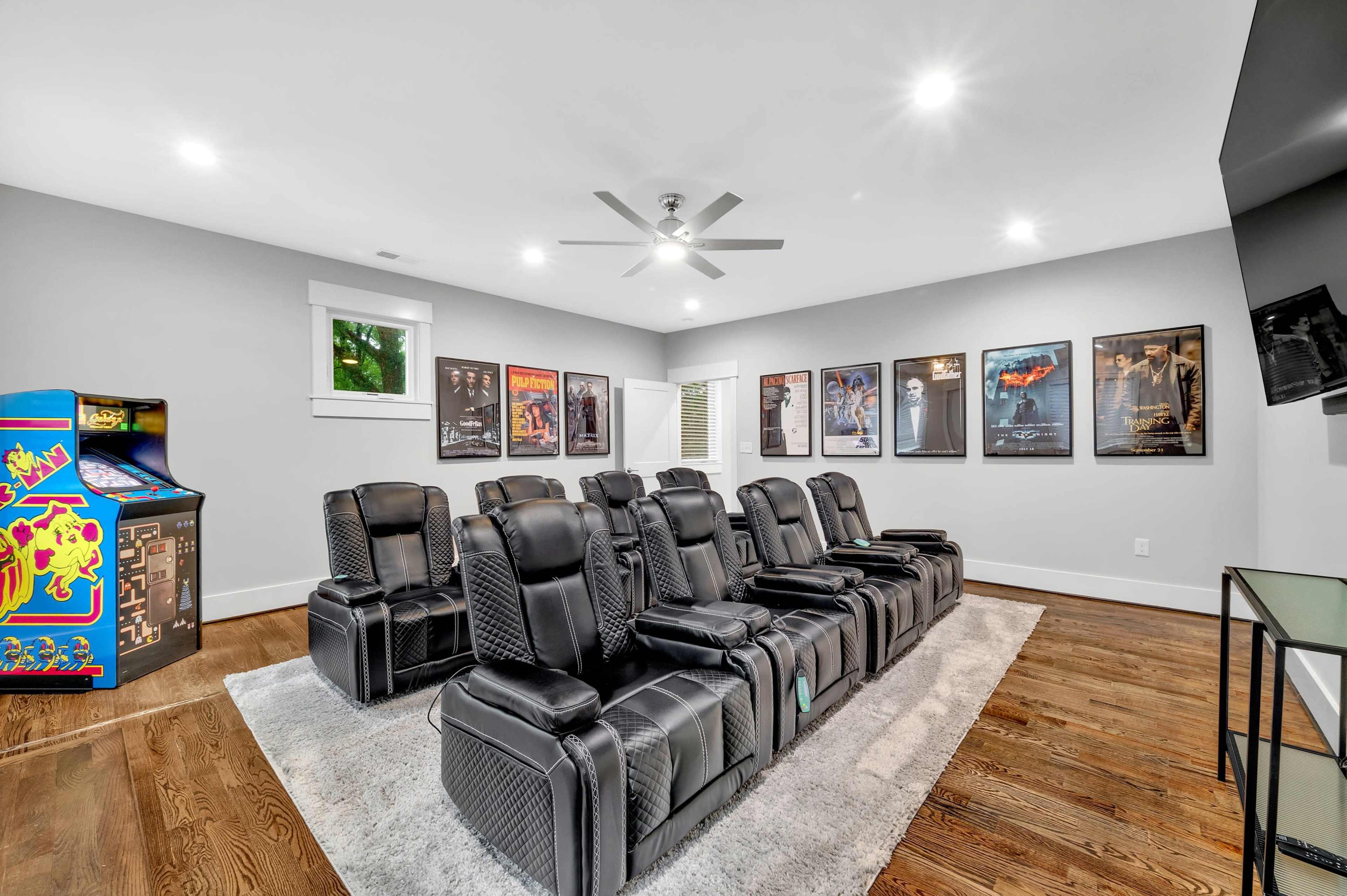 A home theater room with black reclining chairs, a carpeted floor, and movie posters on the walls, alongside a vintage arcade game machine.