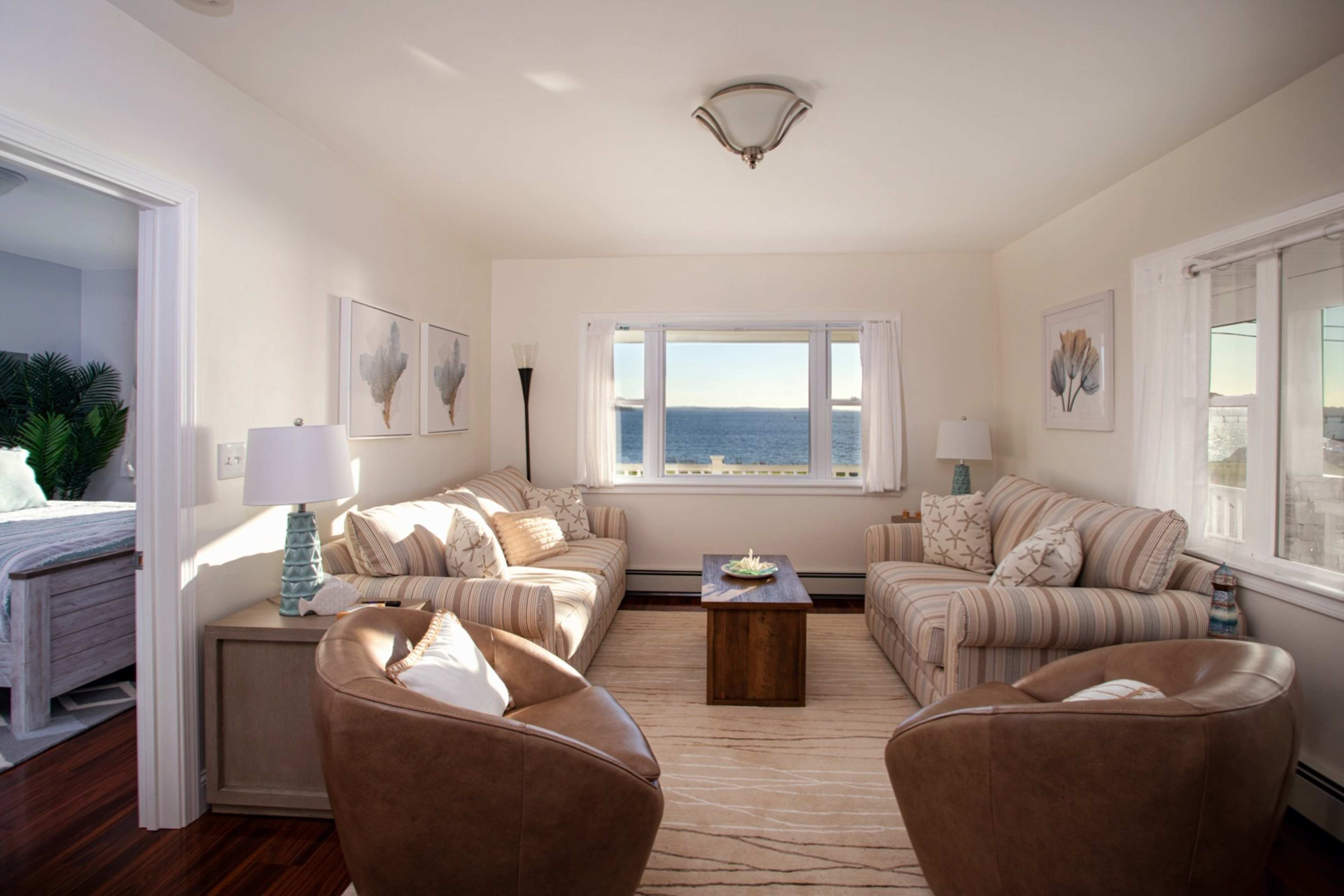 The room features a seating area with two couches and two chairs facing a coffee table, all set against a backdrop of large windows overlooking the ocean.
