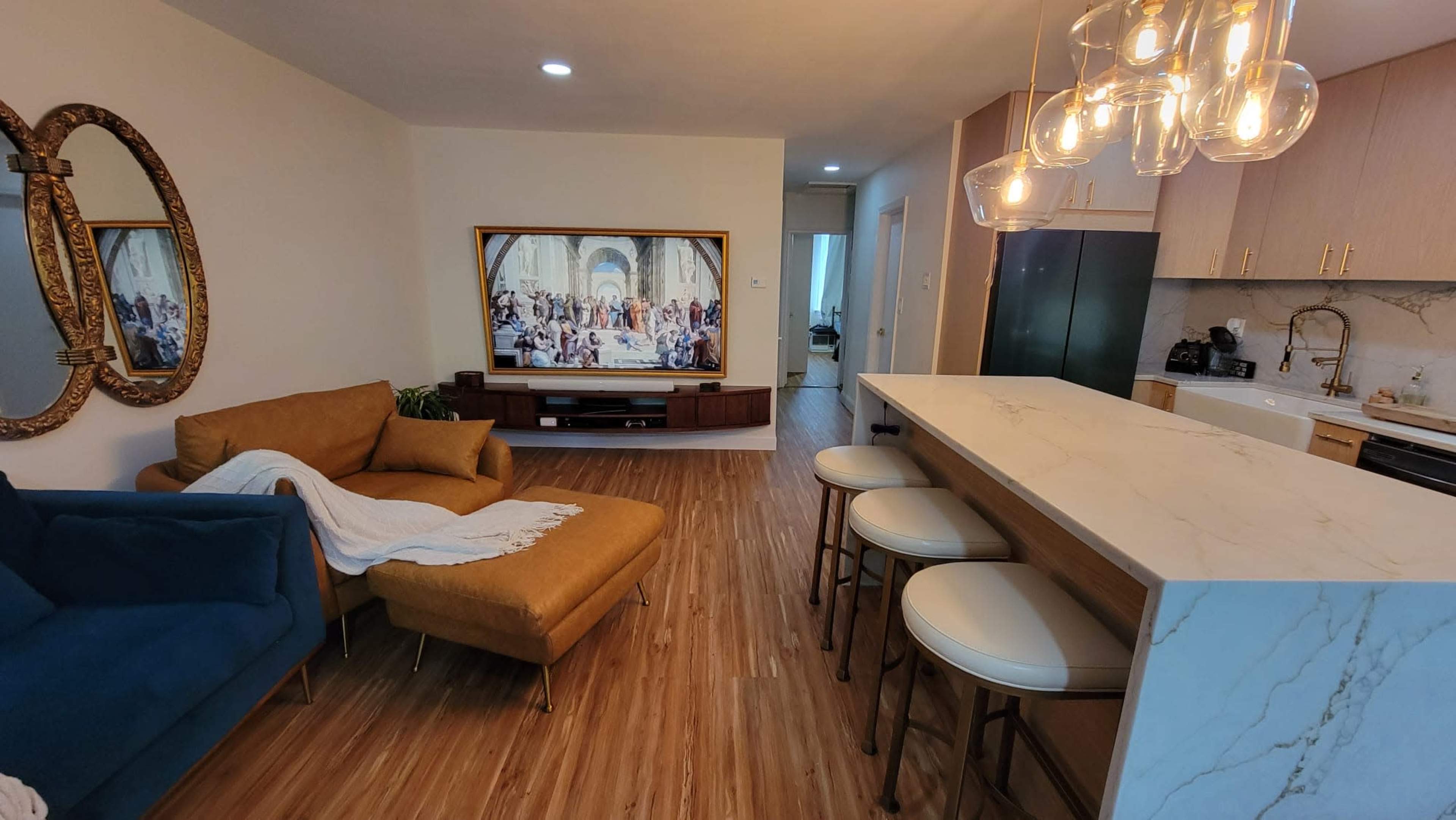 A modern living area featuring a blue sofa, a brown lounge chair, a marble-topped bar with white stools, and a large television displaying a classical painting.