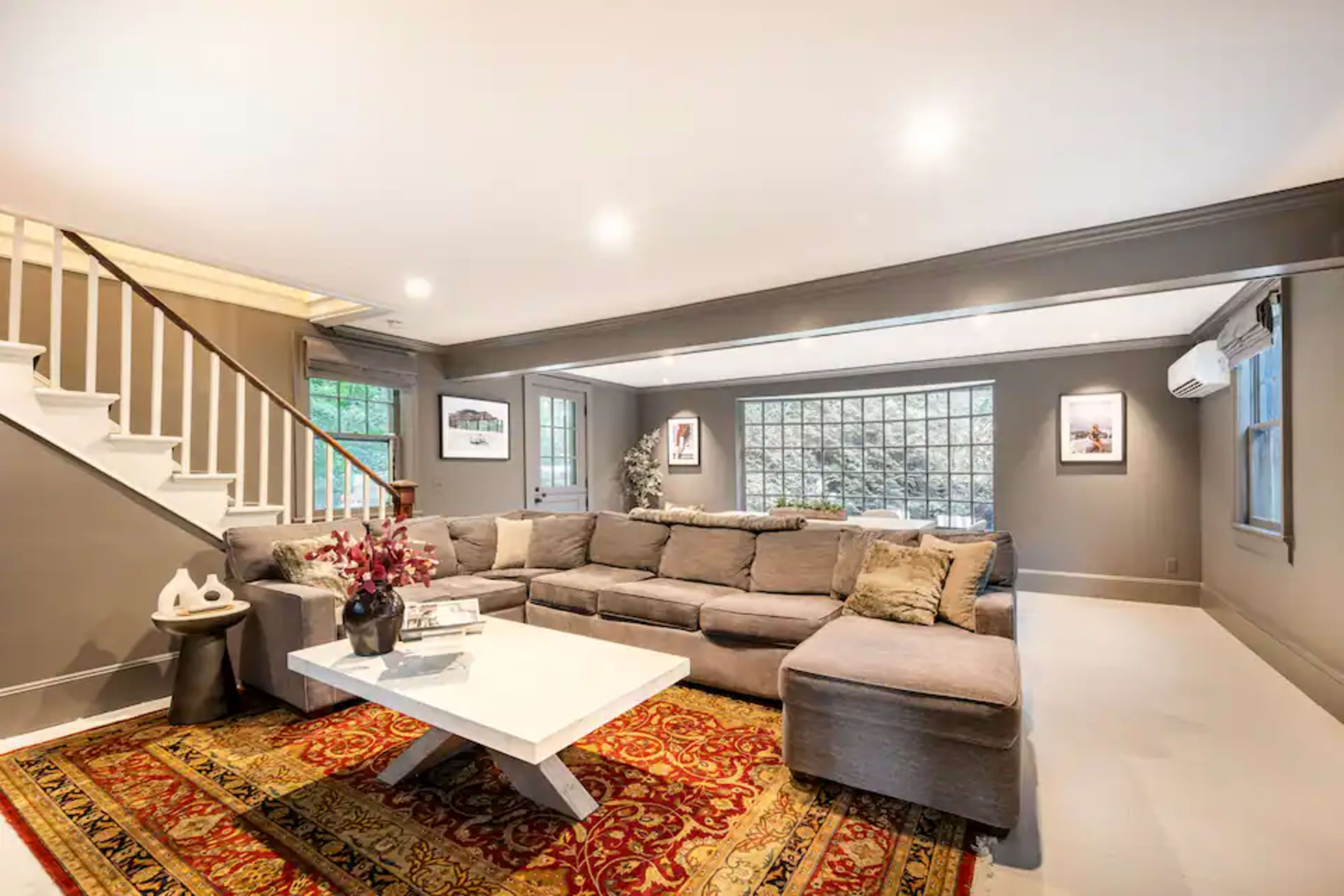 The living room features a large sectional sofa, a white coffee table on a patterned rug, and large windows that allow natural light to enter the space.