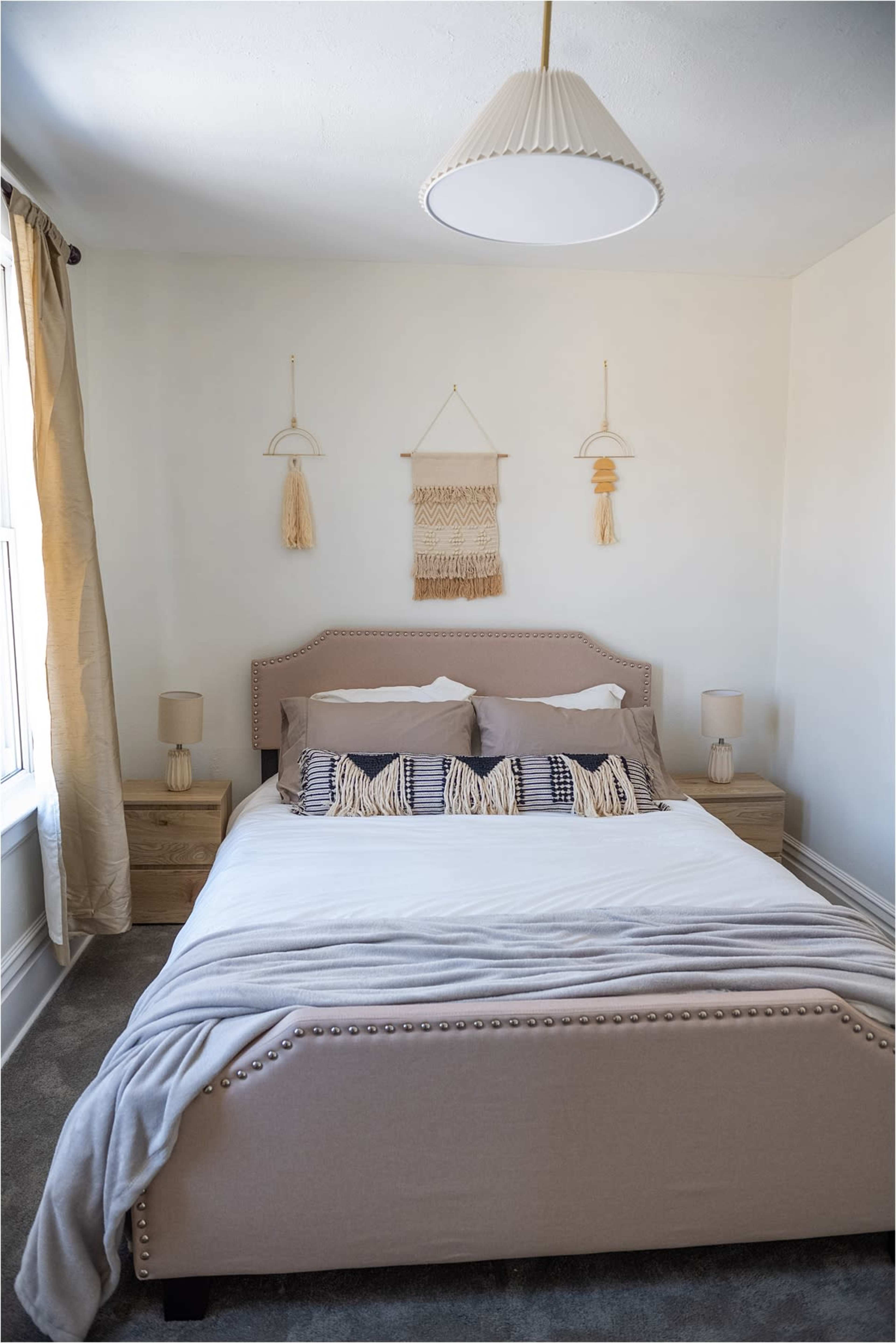 A minimalistic bedroom featuring a beige upholstered bed, two side tables with lamps, and decorative wall hangings.