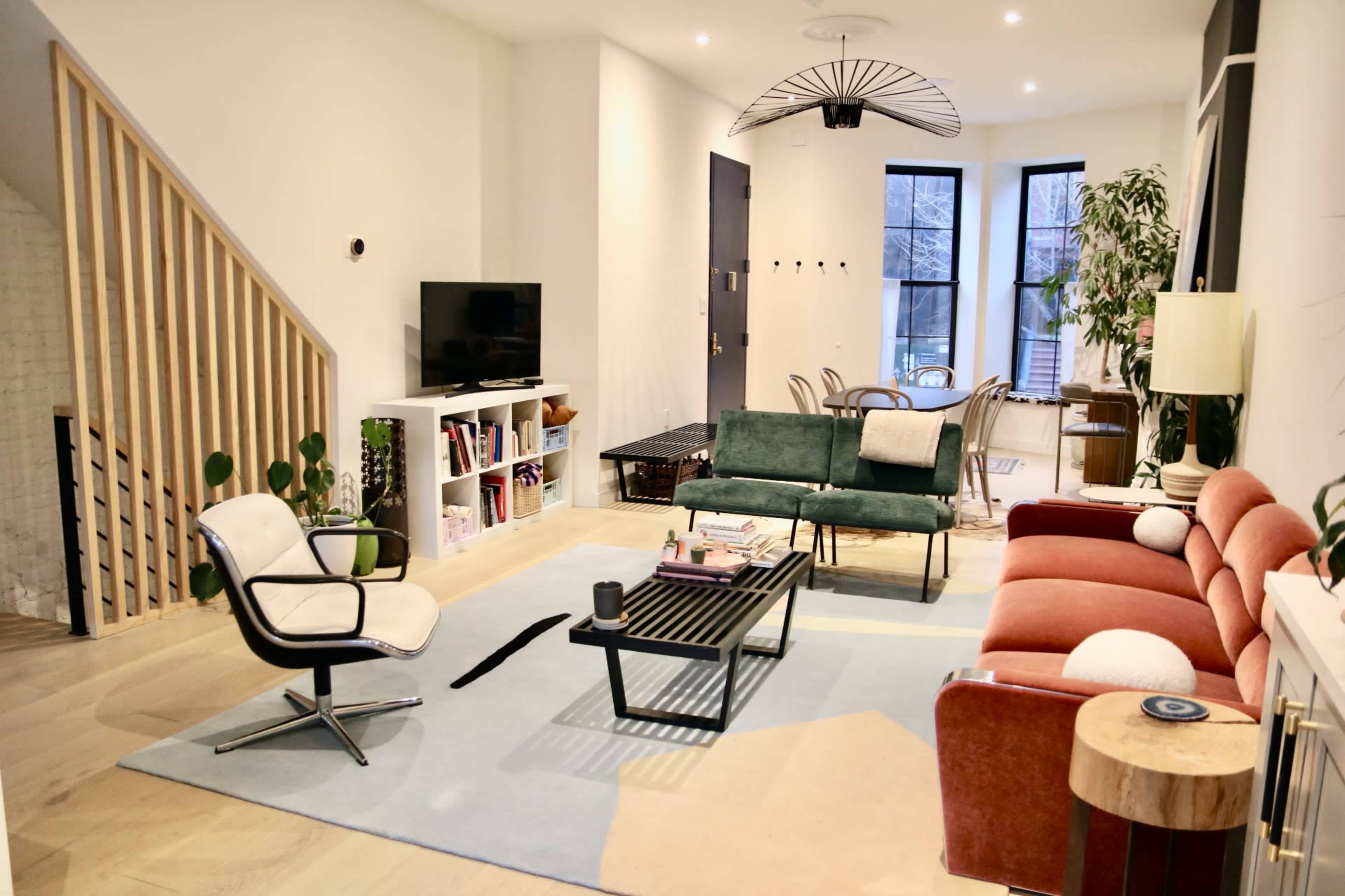 A modern living room features a green sofa, a red couch, and a black coffee table on a light rug, with a staircase in the background and large windows providing natural light.