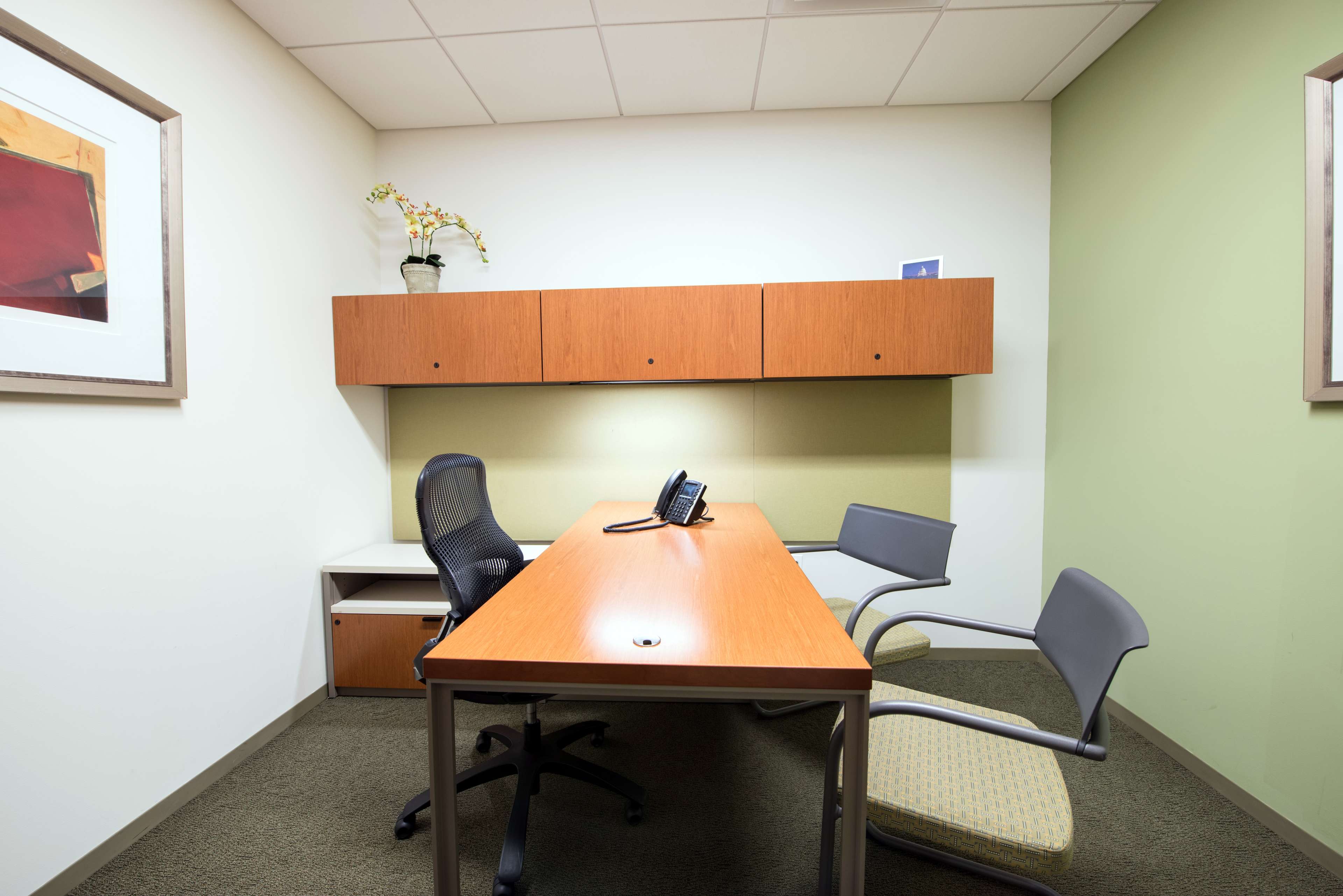 A small office with a wooden desk, two chairs, a phone, and storage cabinets mounted above the desk.