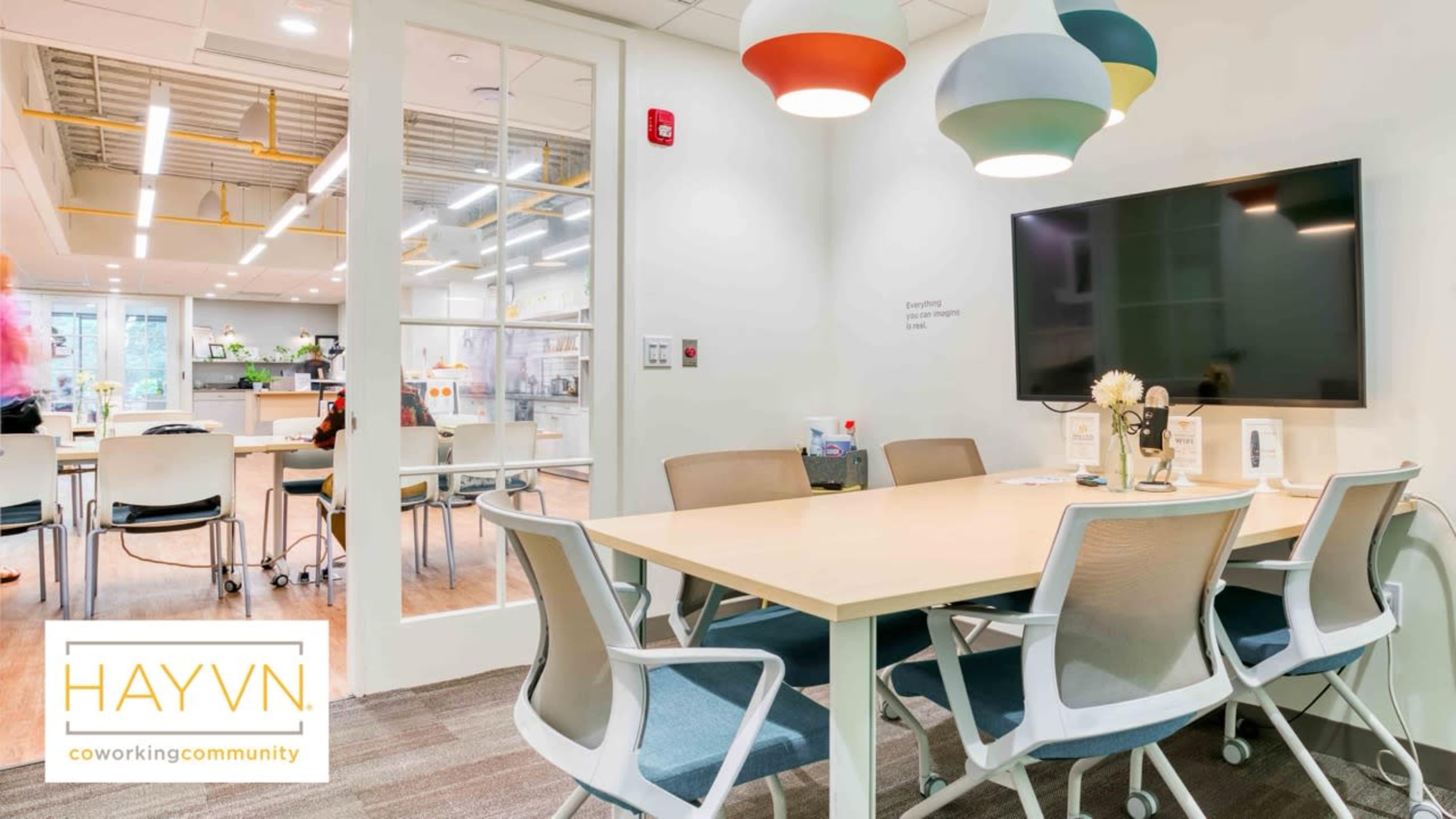 The image shows a modern coworking space featuring a meeting room with a large table, several chairs, and a wall-mounted TV, along with colorful pendant lights overhead.