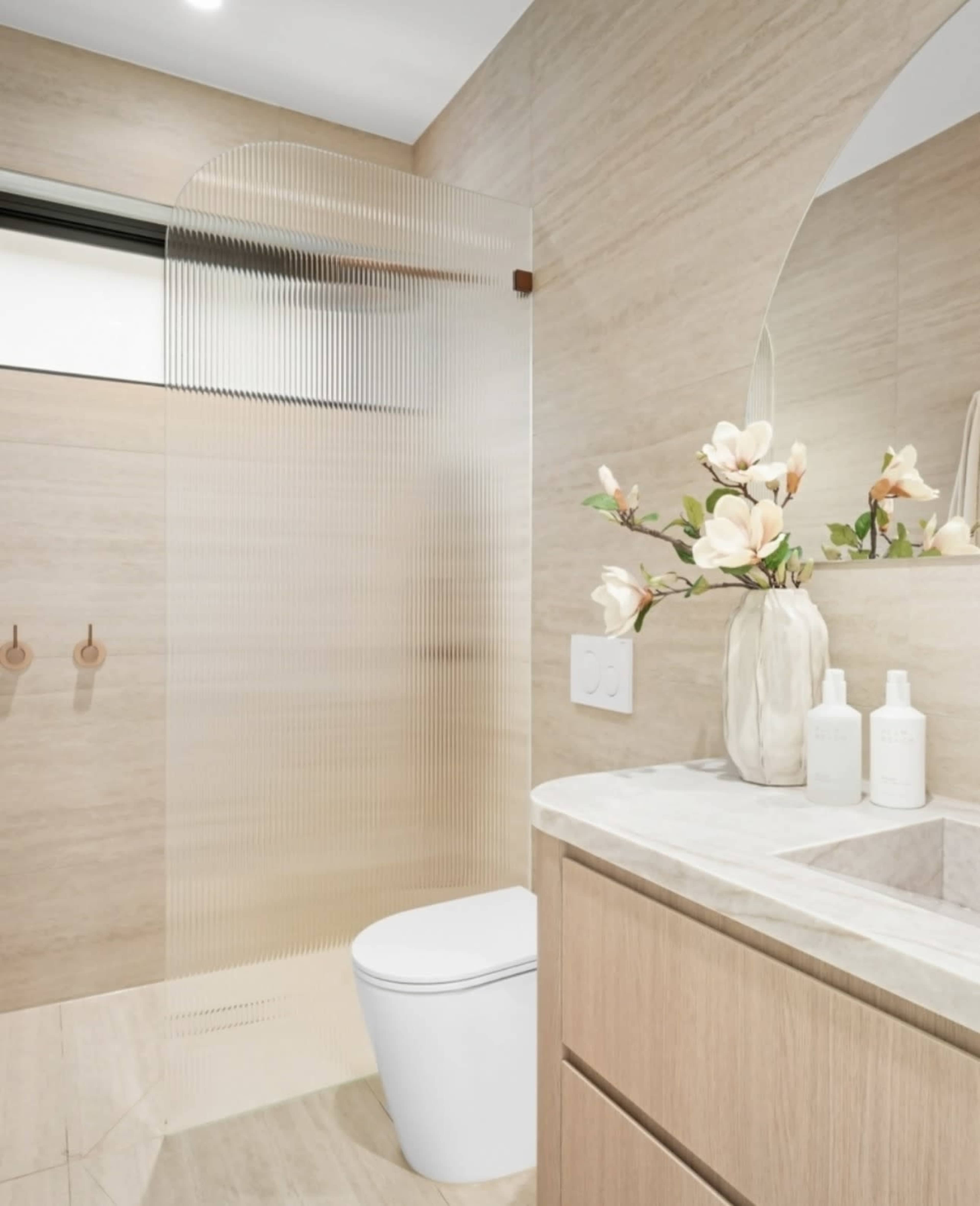 The image shows a modern bathroom featuring a glass shower partition, a white toilet, a wooden vanity with a marble countertop, and decorative flowers.