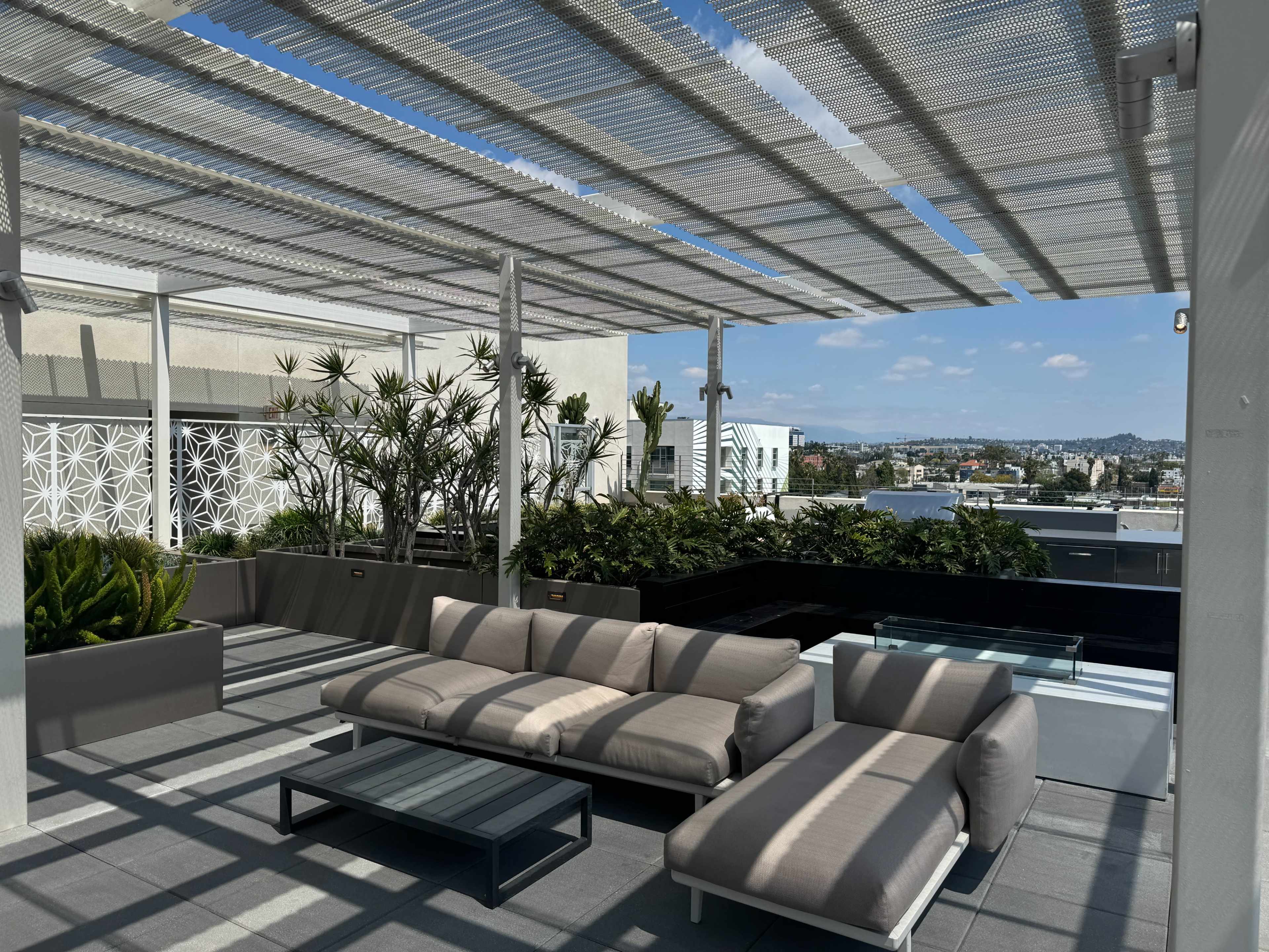 A modern rooftop lounge area features a couch and coffee table under a patterned pergola, surrounded by potted plants and a cityscape view.