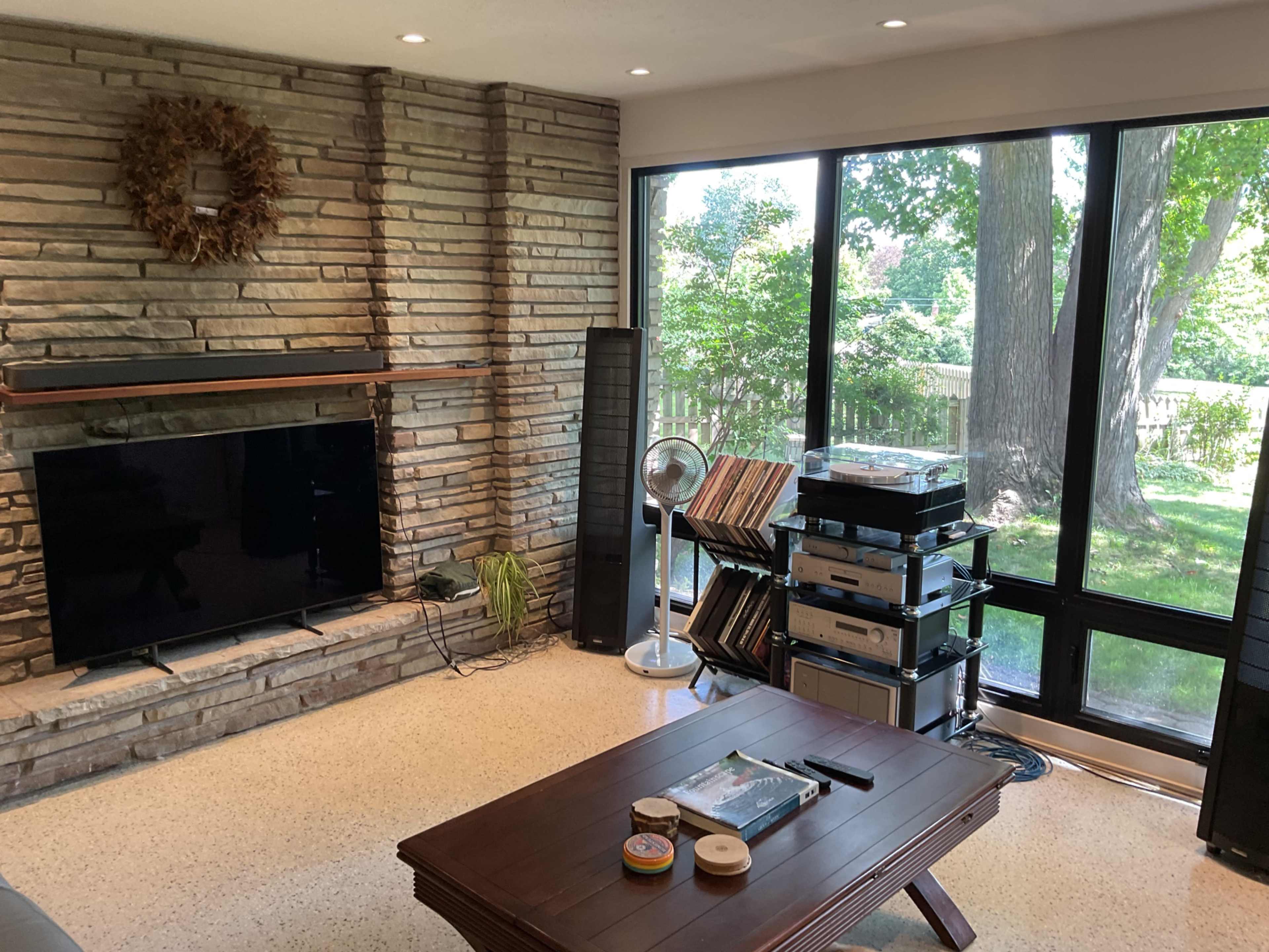 The living room features a stone wall with a television, a coffee table, and a set of audio equipment, alongside large windows showing a green yard and trees.