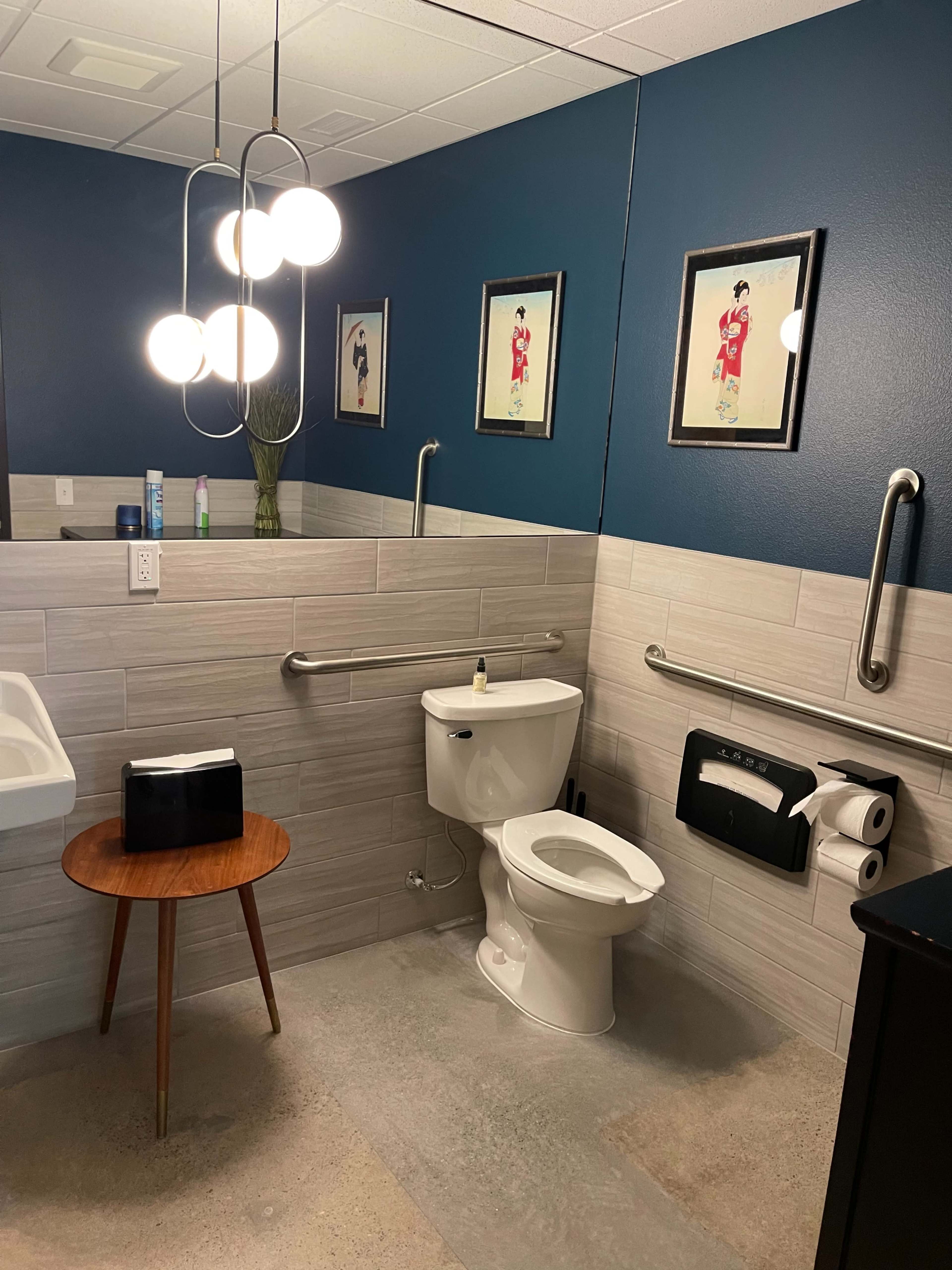 The bathroom features a white toilet, a sink on the left, a wooden table, and mirrors with framed artwork on a blue wall.