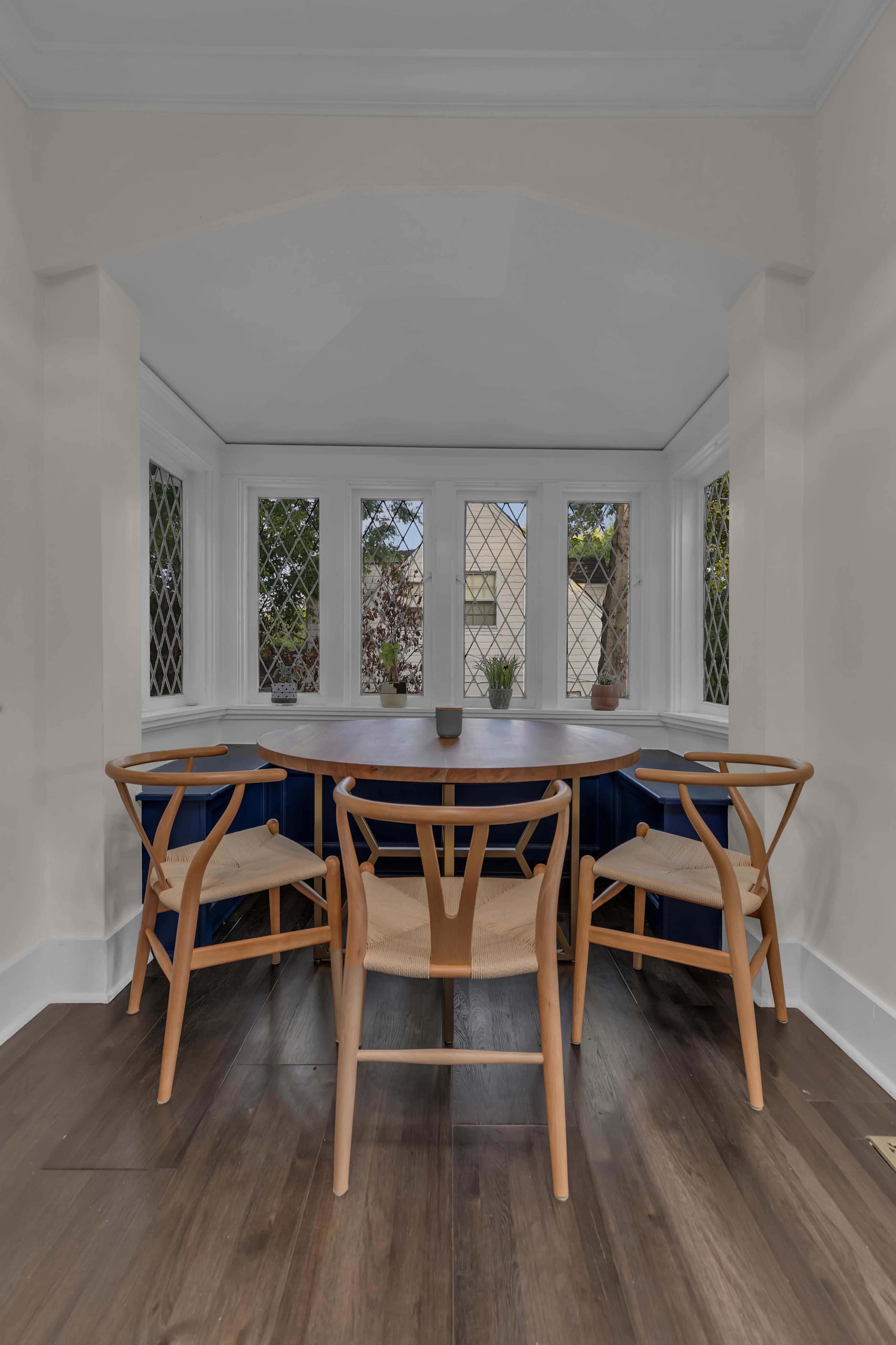 A corner dining nook features a round table surrounded by four wooden chairs, situated near large windows with diamond-patterned panes.