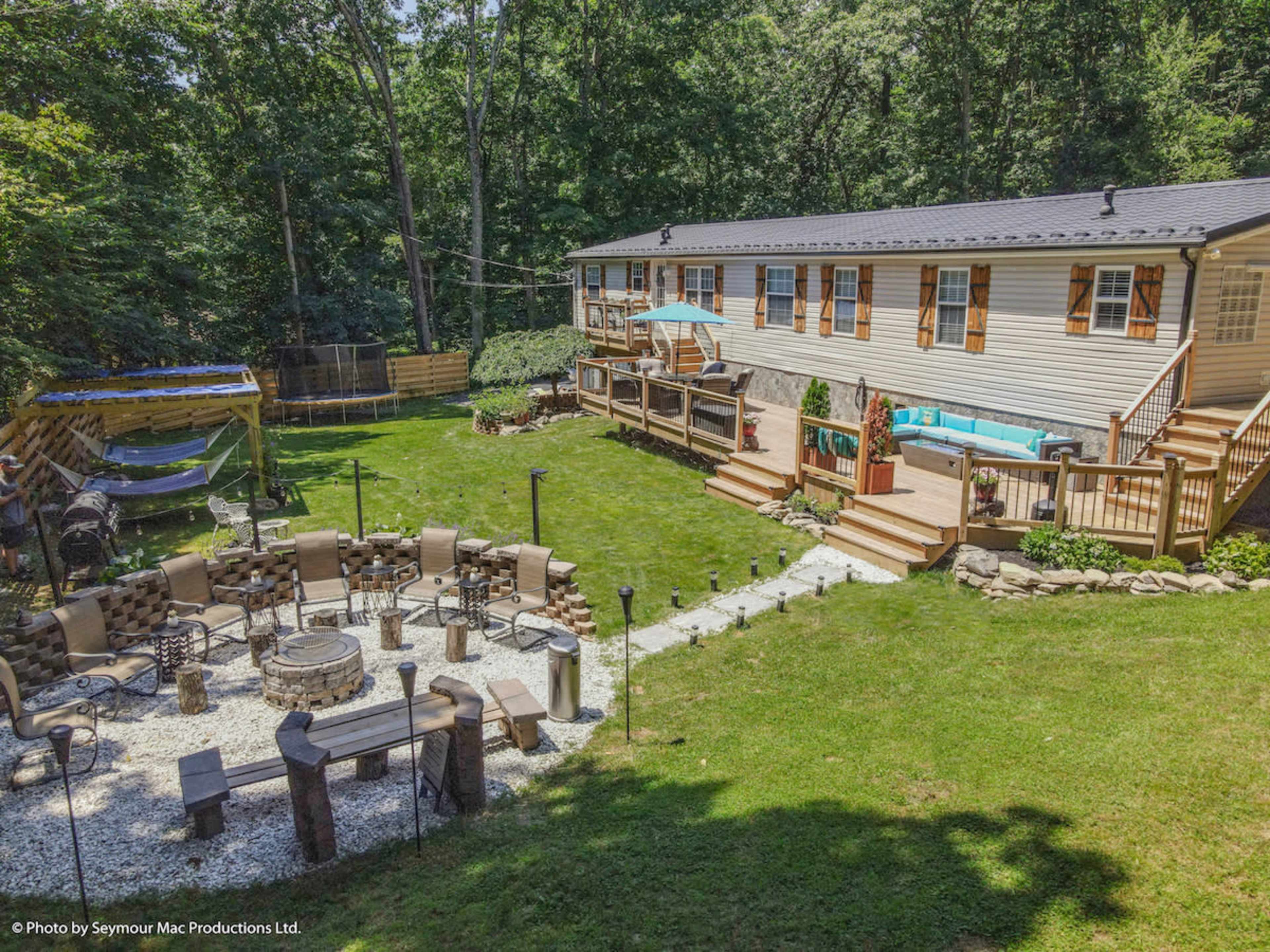 A landscaped backyard featuring a large wooden deck, a stone fire pit area surrounded by seating, and a well-maintained lawn, along with a house set against a wooded backdrop.
