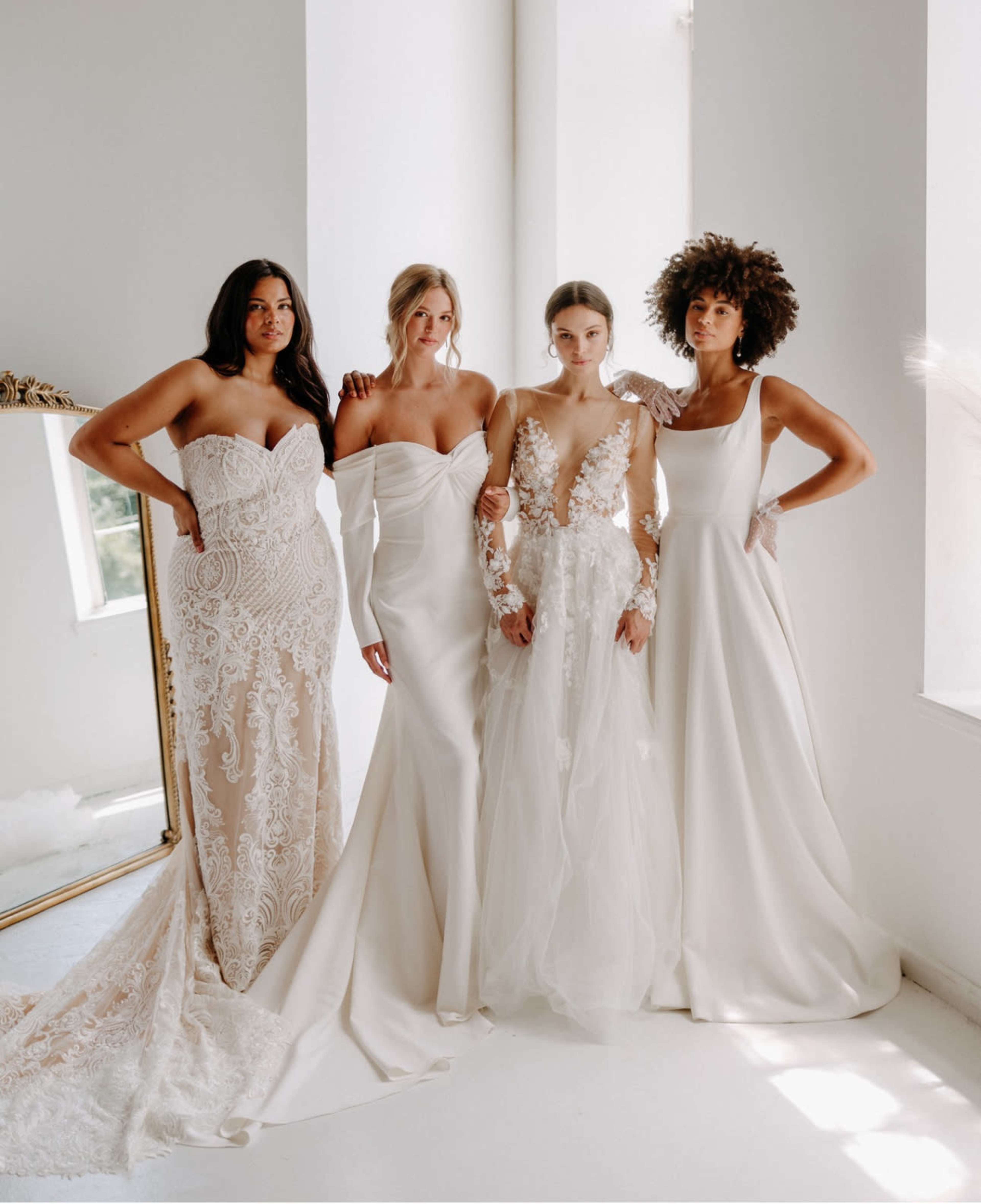 Four women stand together in elegant wedding dresses, showcasing a variety of styles and designs against a bright, minimalist backdrop.