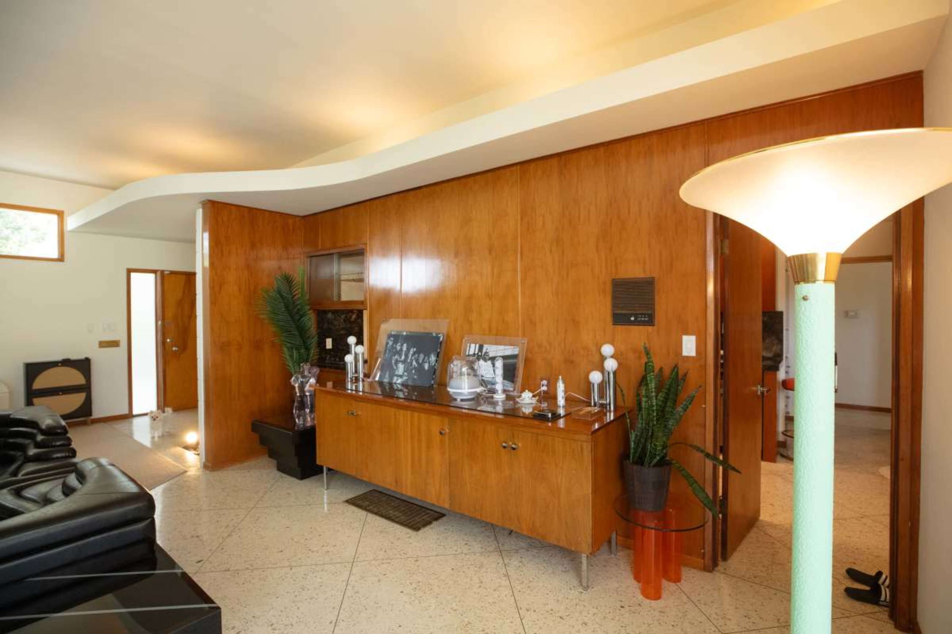 The image shows a mid-century modern living room featuring a wooden sideboard, a floor lamp, and large black leather sofas.