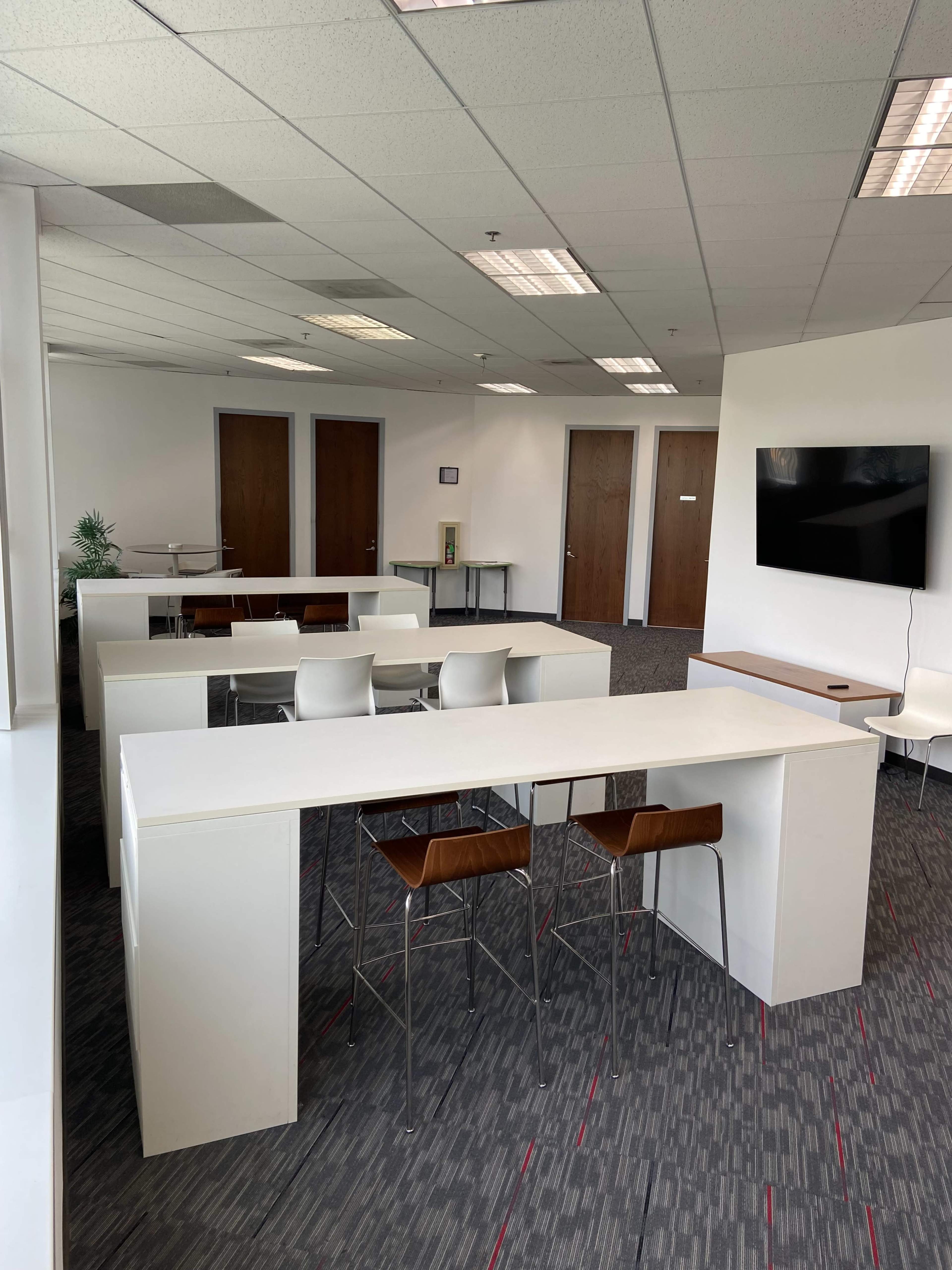 A modern office space with white tables and chairs arranged neatly, a large TV on the wall, and wooden doors in the background.