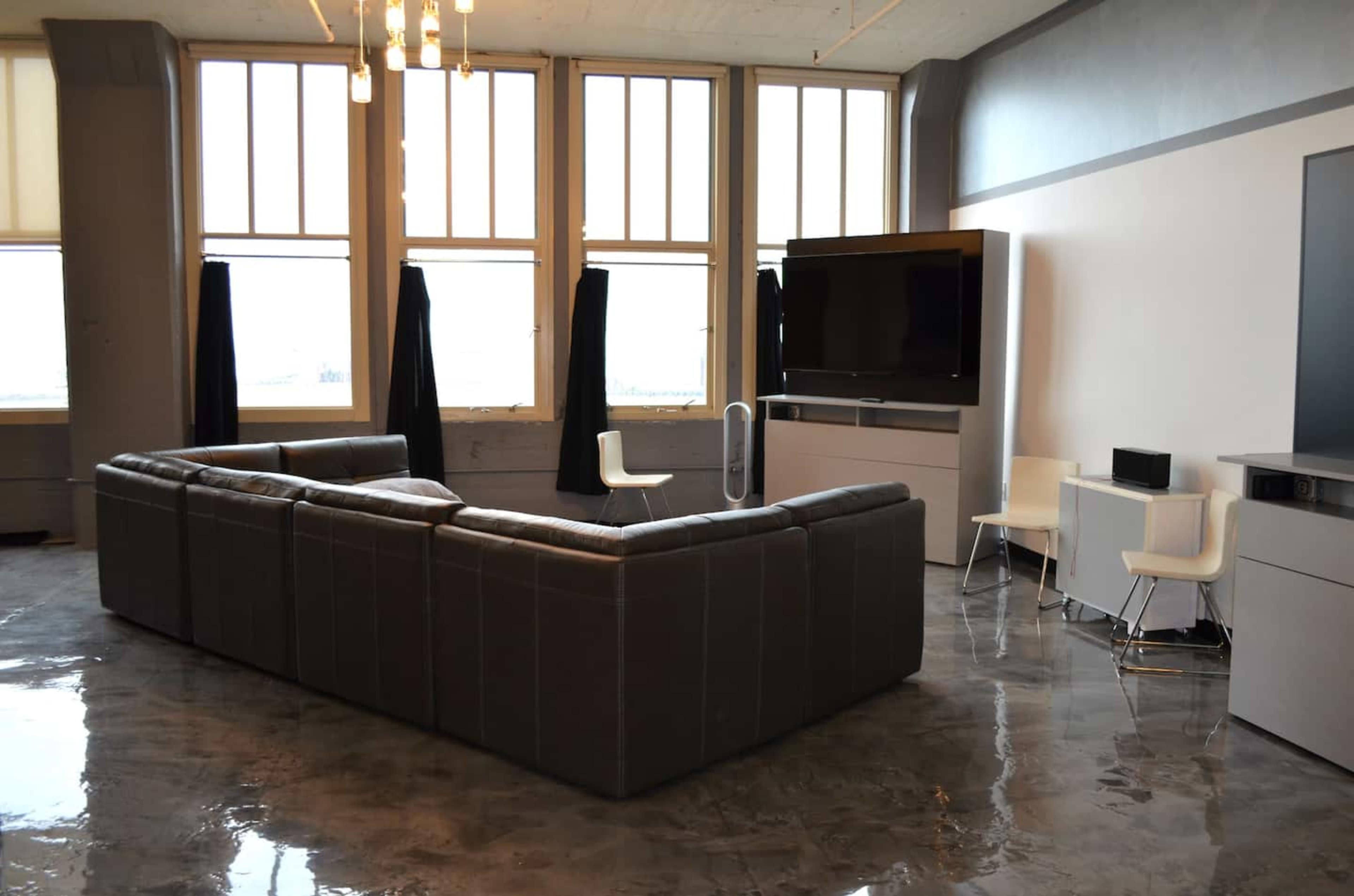 A spacious living room features a large sectional sofa, a flat-screen television, and modern furnishings with large windows allowing natural light.