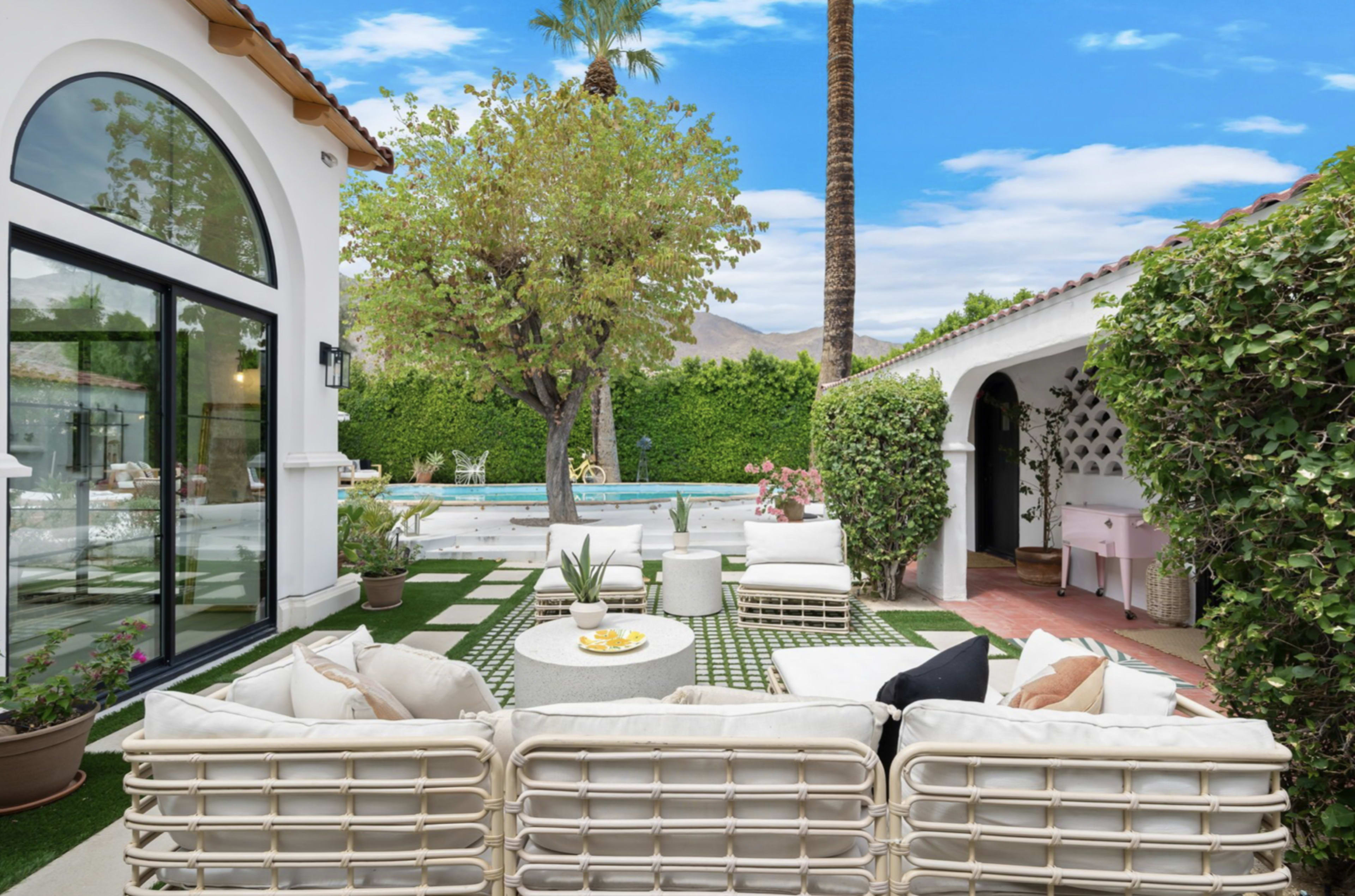 The image shows a backyard area with a seating arrangement, surrounded by greenery, a pool, and a clear blue sky.