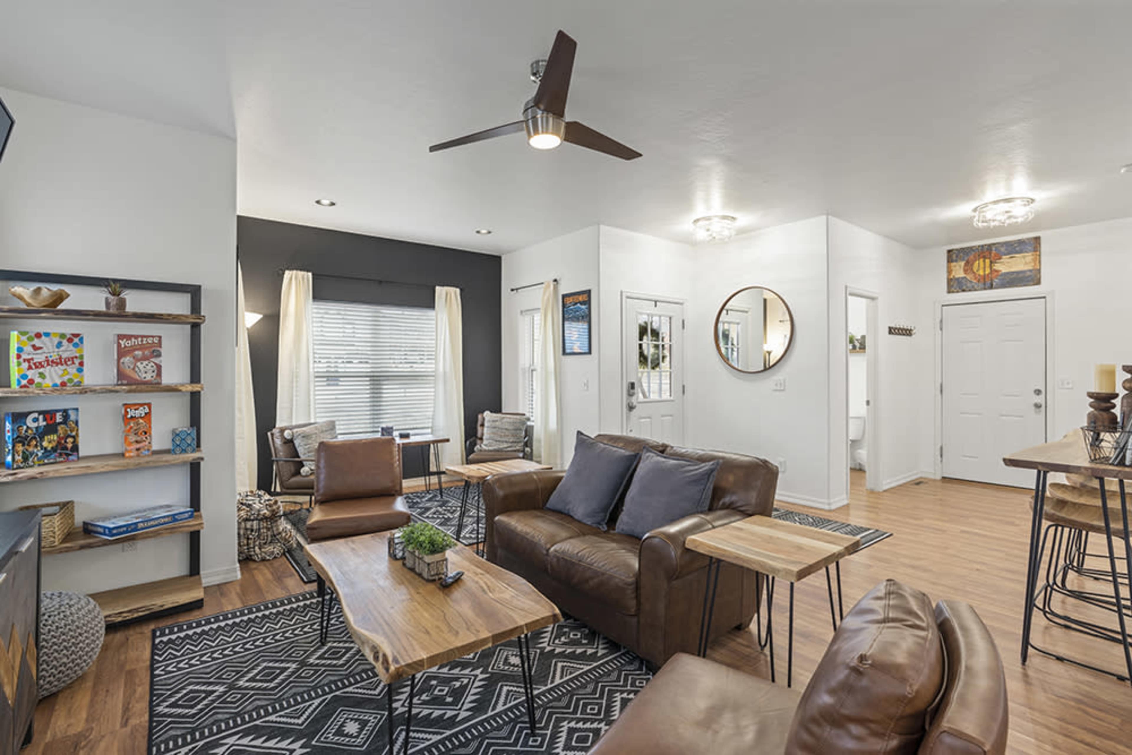A modern living room featuring brown leather furniture, a wooden coffee table, and a patterned rug, with a ceiling fan and large windows.