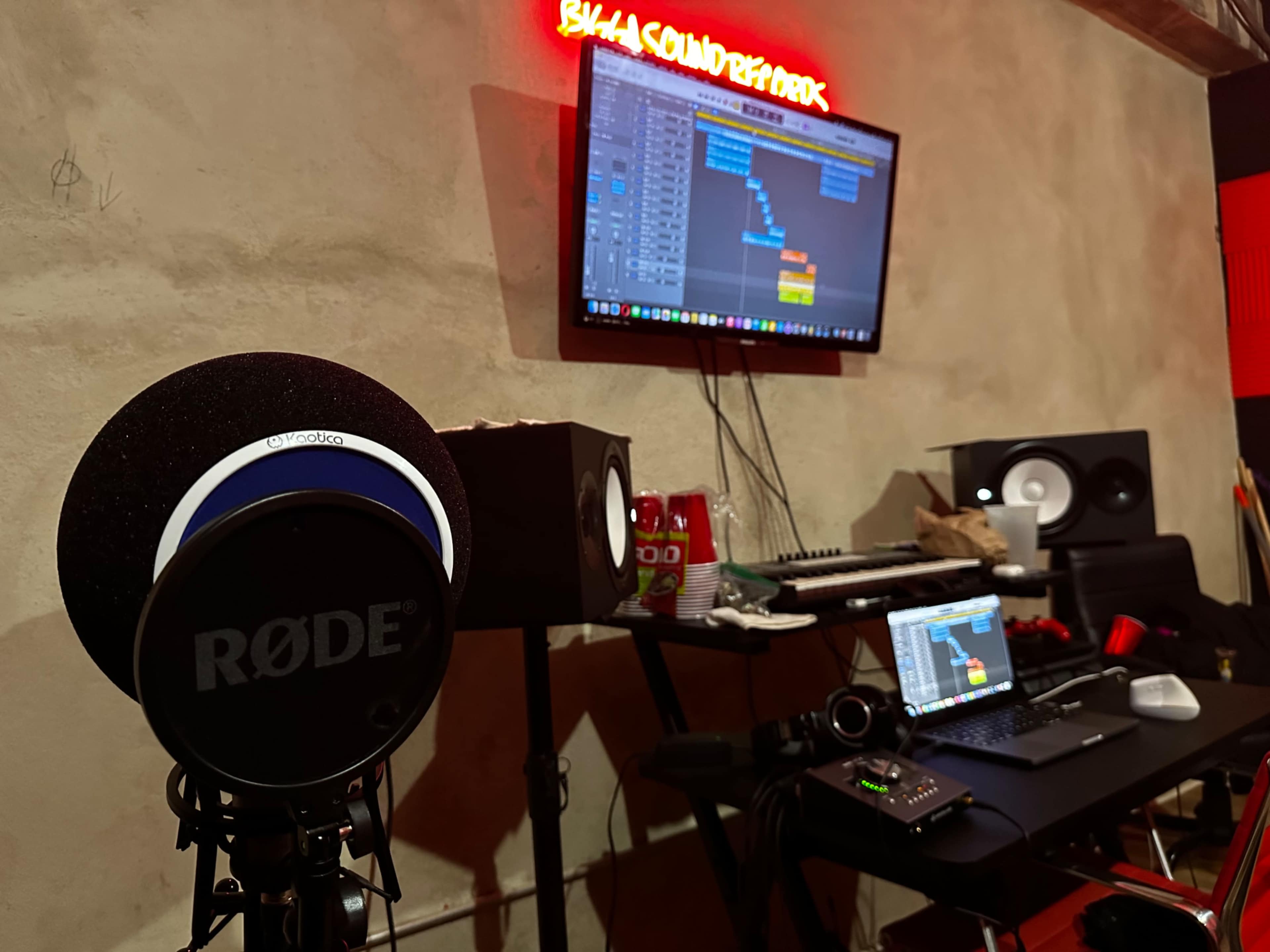 A microphone stands in the foreground, while a computer screen displays audio editing software alongside speakers and various equipment on a desk.