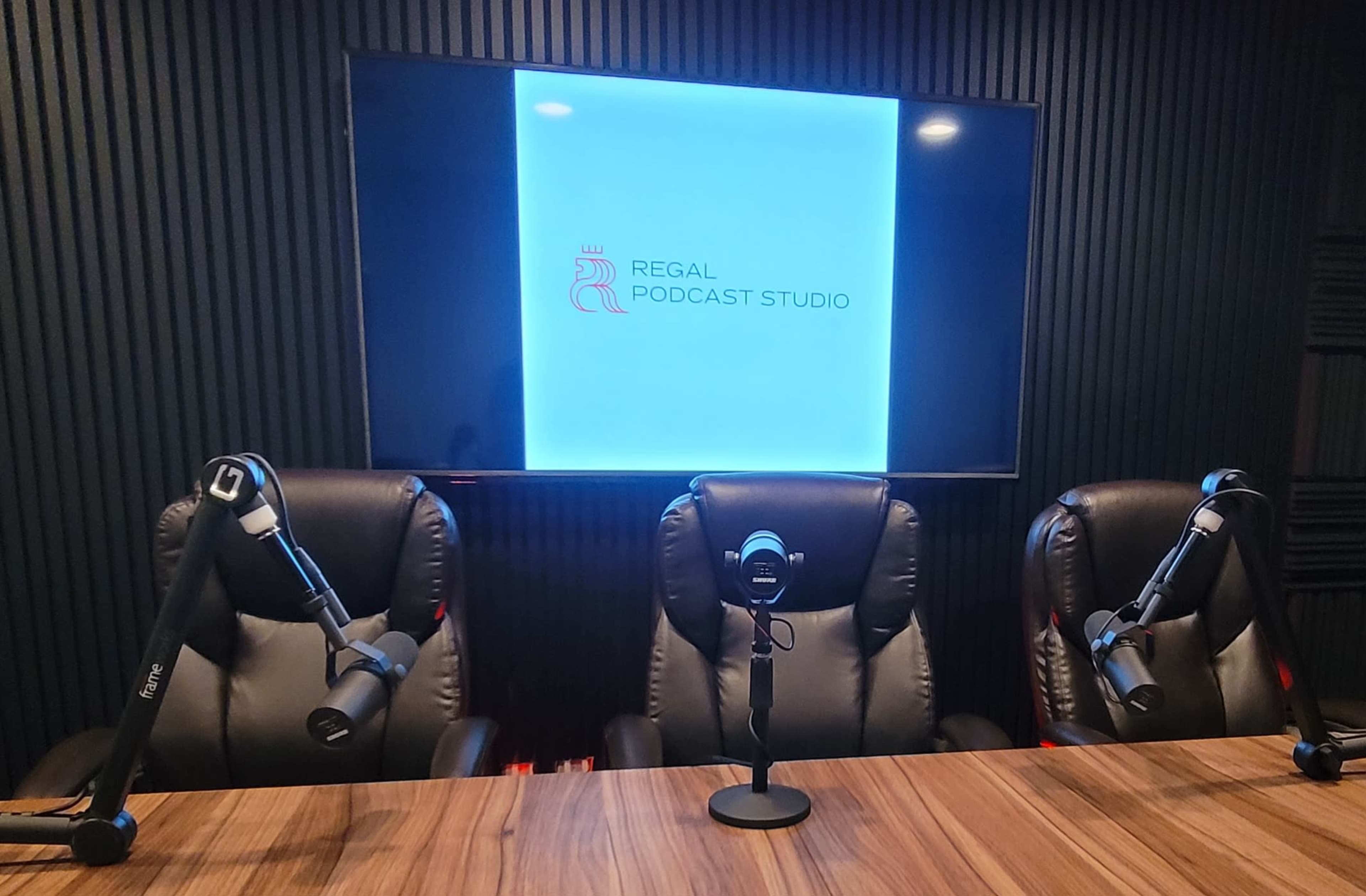 A podcast studio setup featuring three black chairs with microphone stands, a wooden table, and a large screen displaying the text "Regal Podcast Studio."