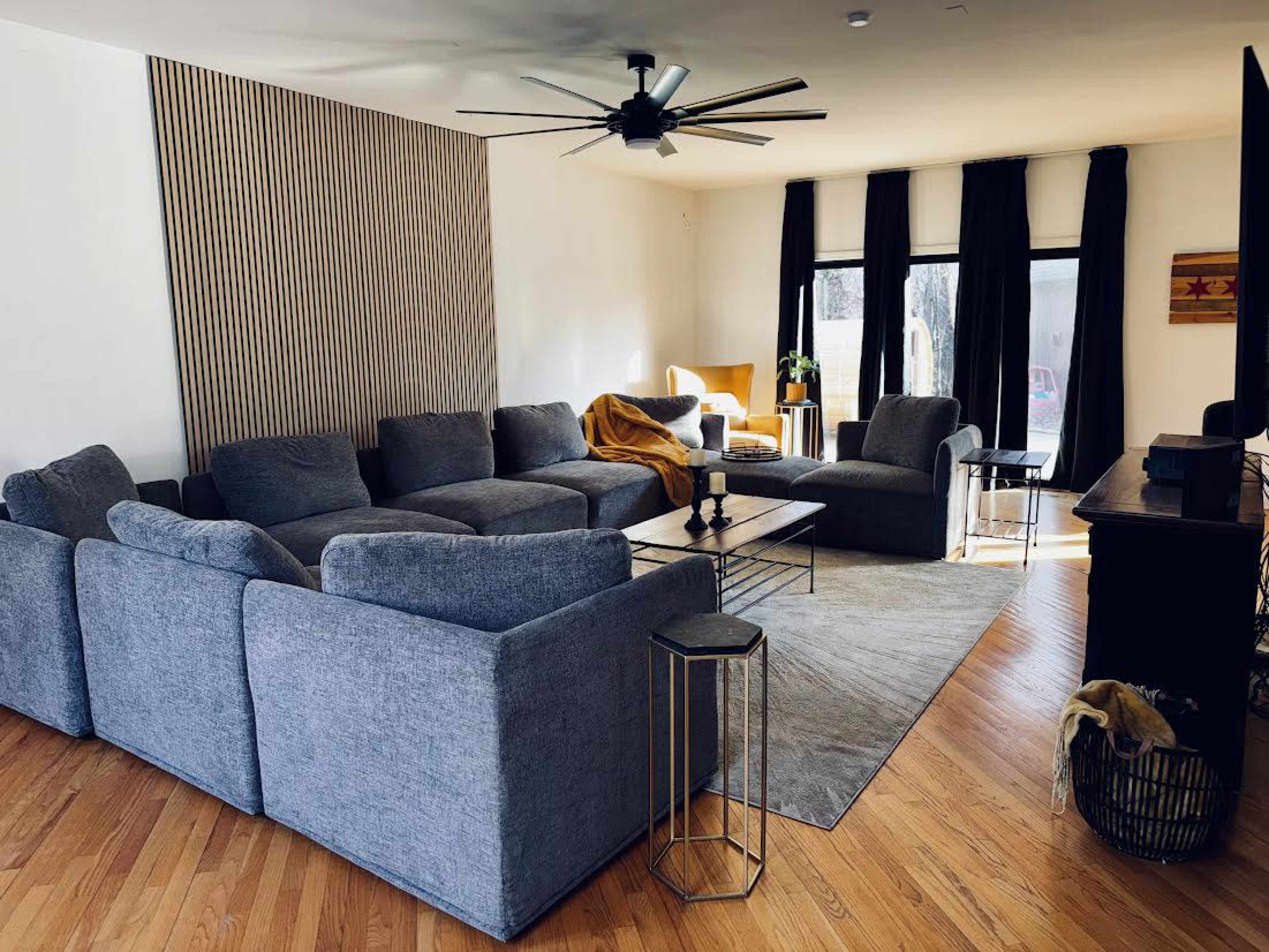 The living room features a large gray sectional couch, a coffee table, and a warm throw draped over a chair, with large windows allowing natural light to flow in.